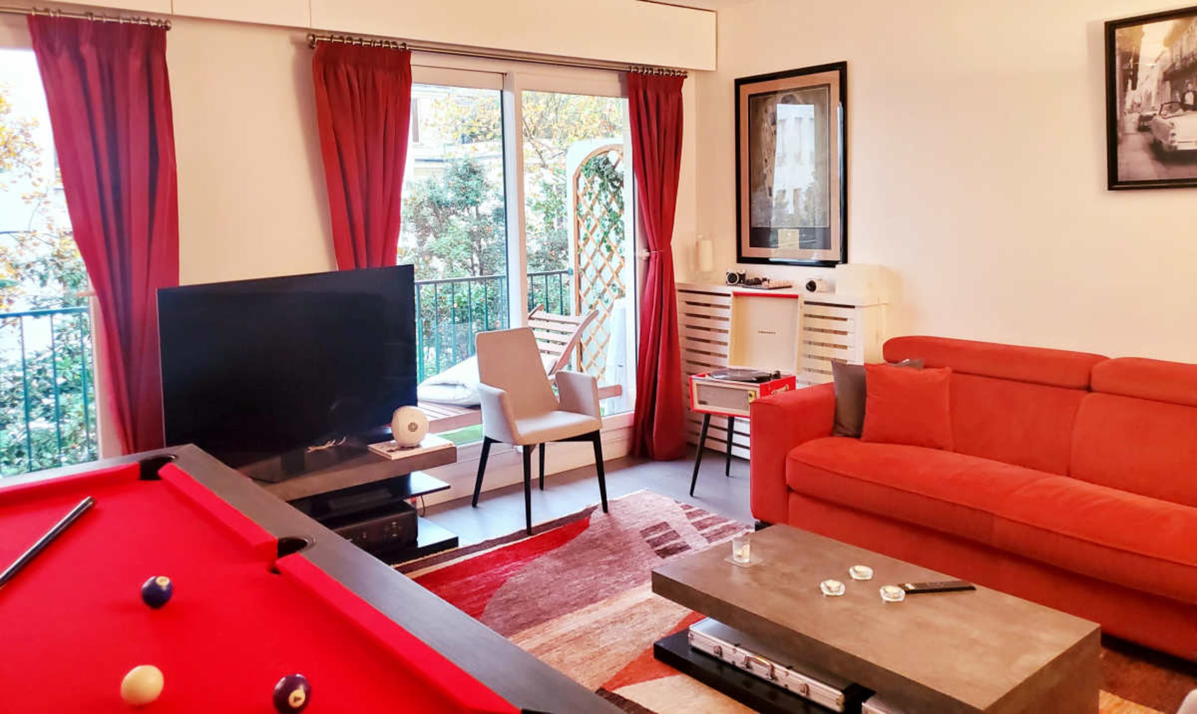 The image depicts a living room featuring a red sofa, a pool table, a television, and a balcony with outdoor greenery visible through large windows.