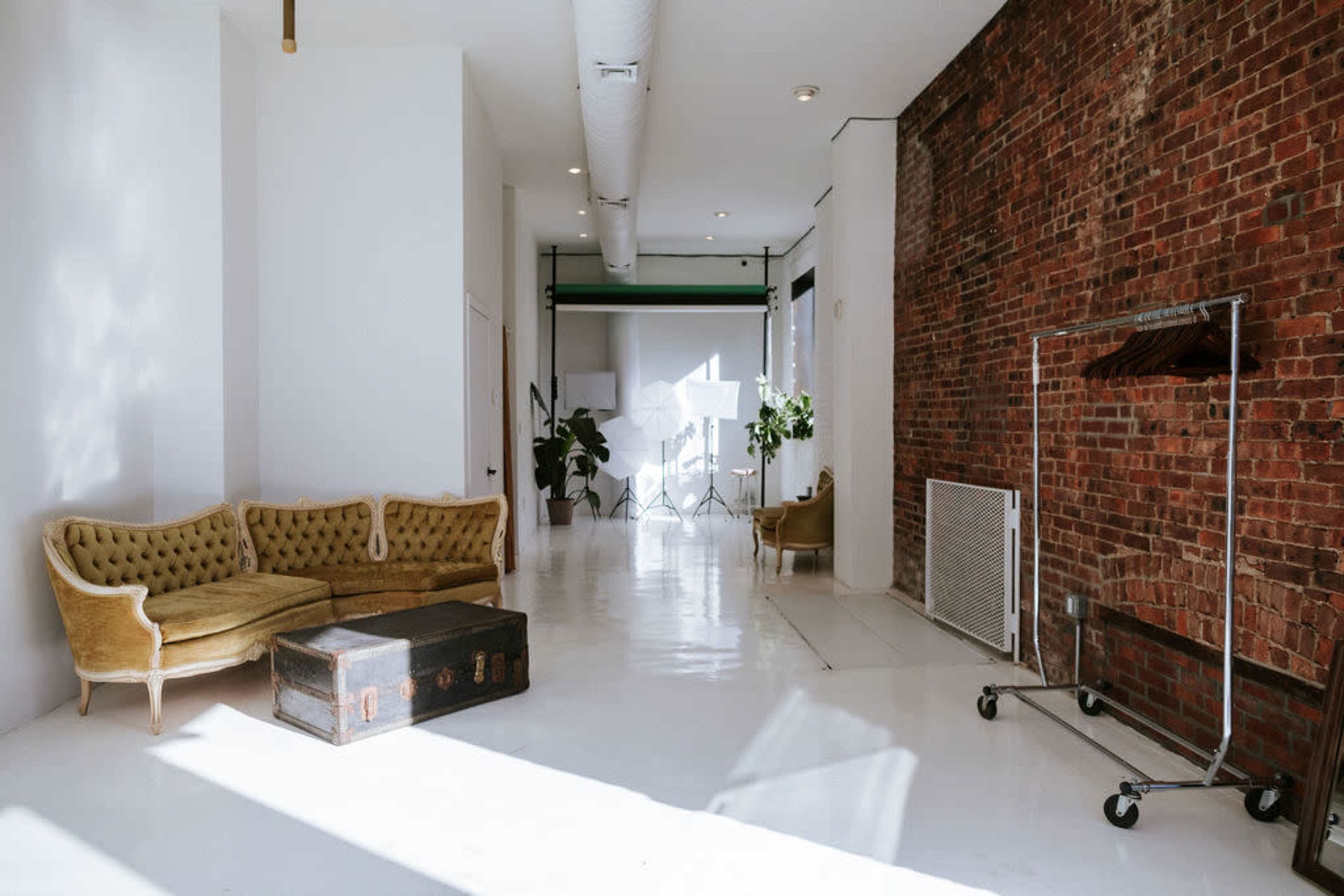 The image shows a spacious interior with a white floor, a vintage yellow couch, a wooden trunk, a clothing rack, and potted plants against a brick wall.
