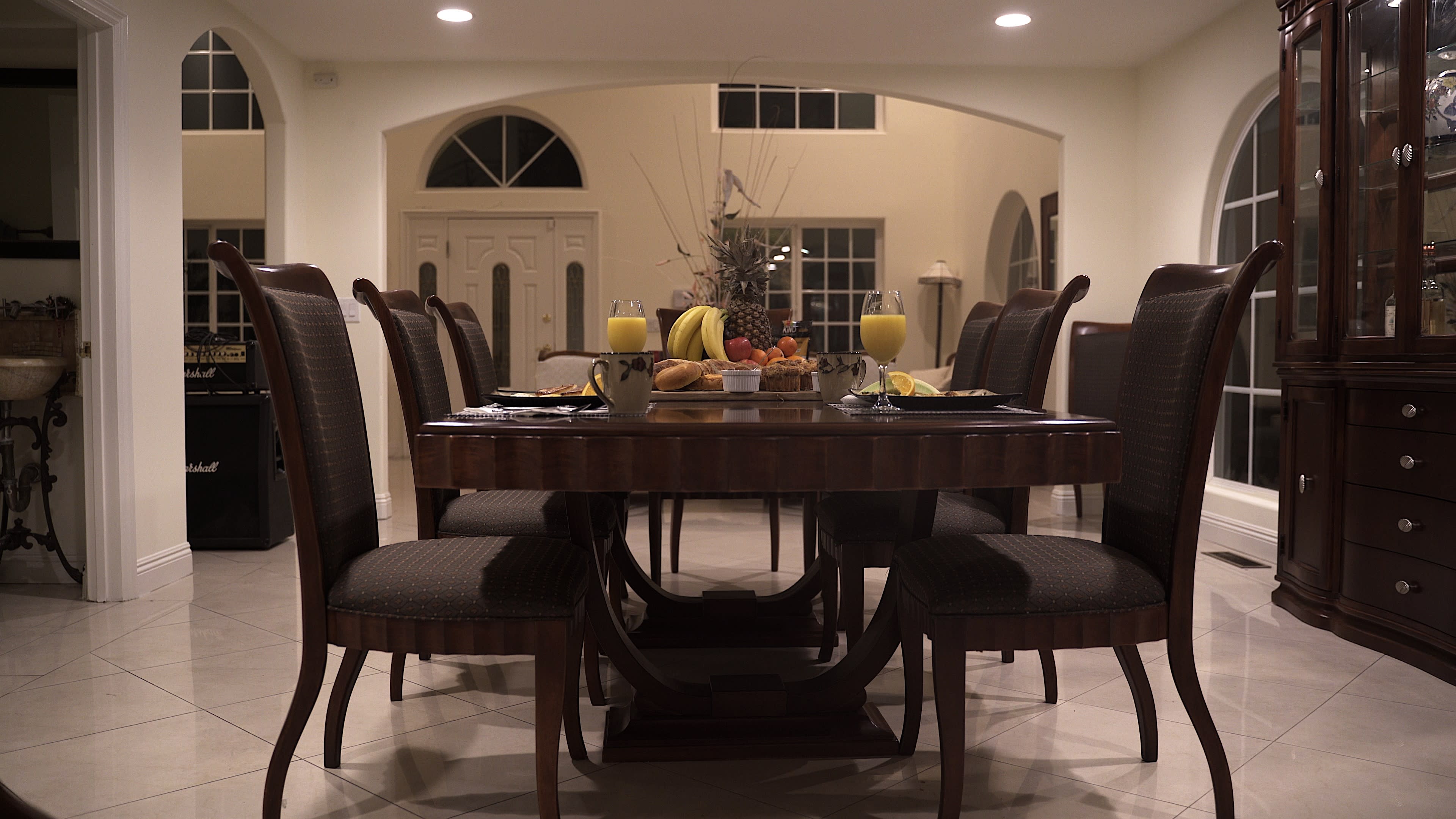 A dining table is set with fruits and beverages, surrounded by chairs in a well-lit room with large windows.