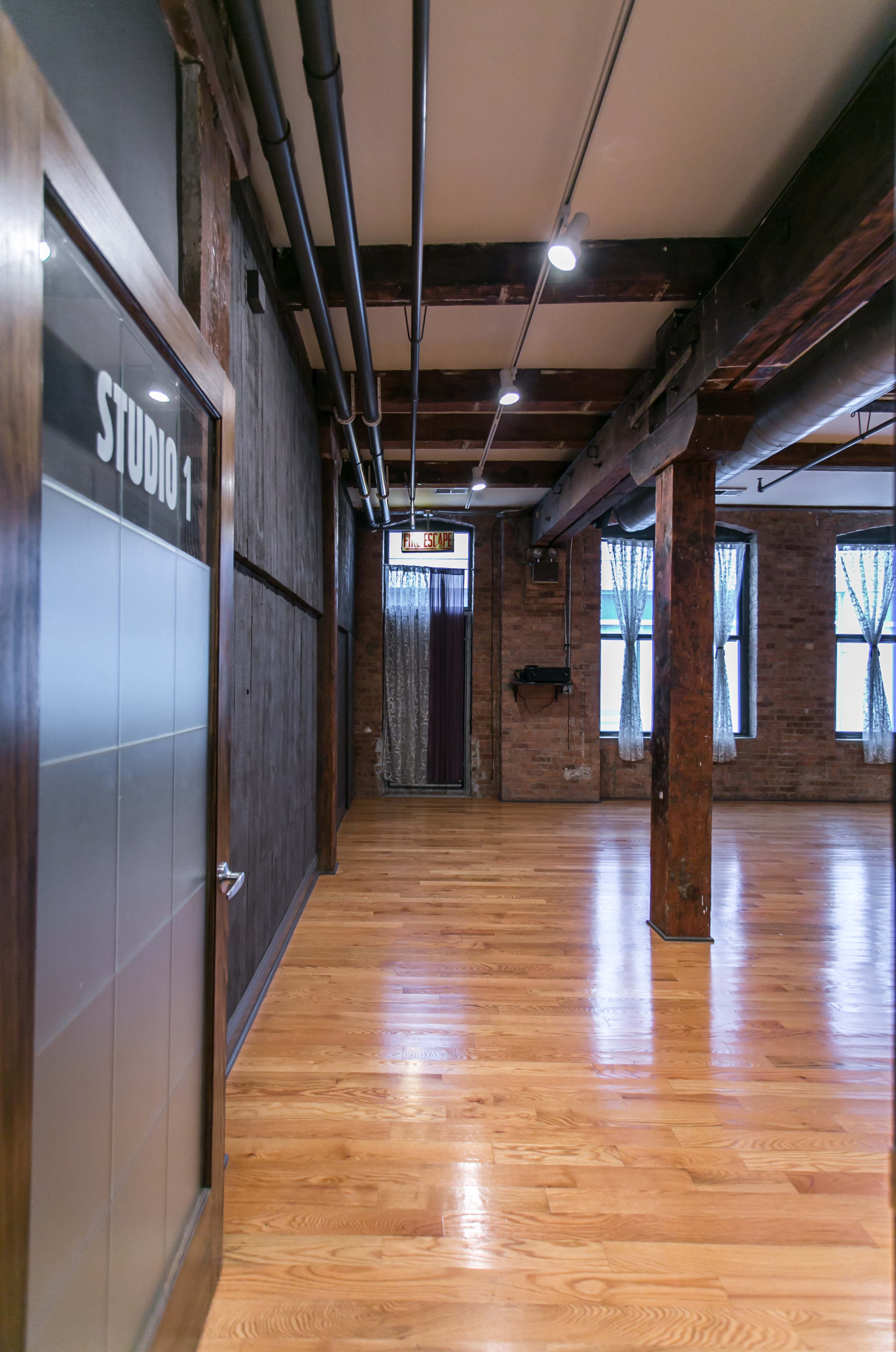 A spacious interior hallway leading to "Studio 1," with wooden beams and floor, and large windows at the far end.