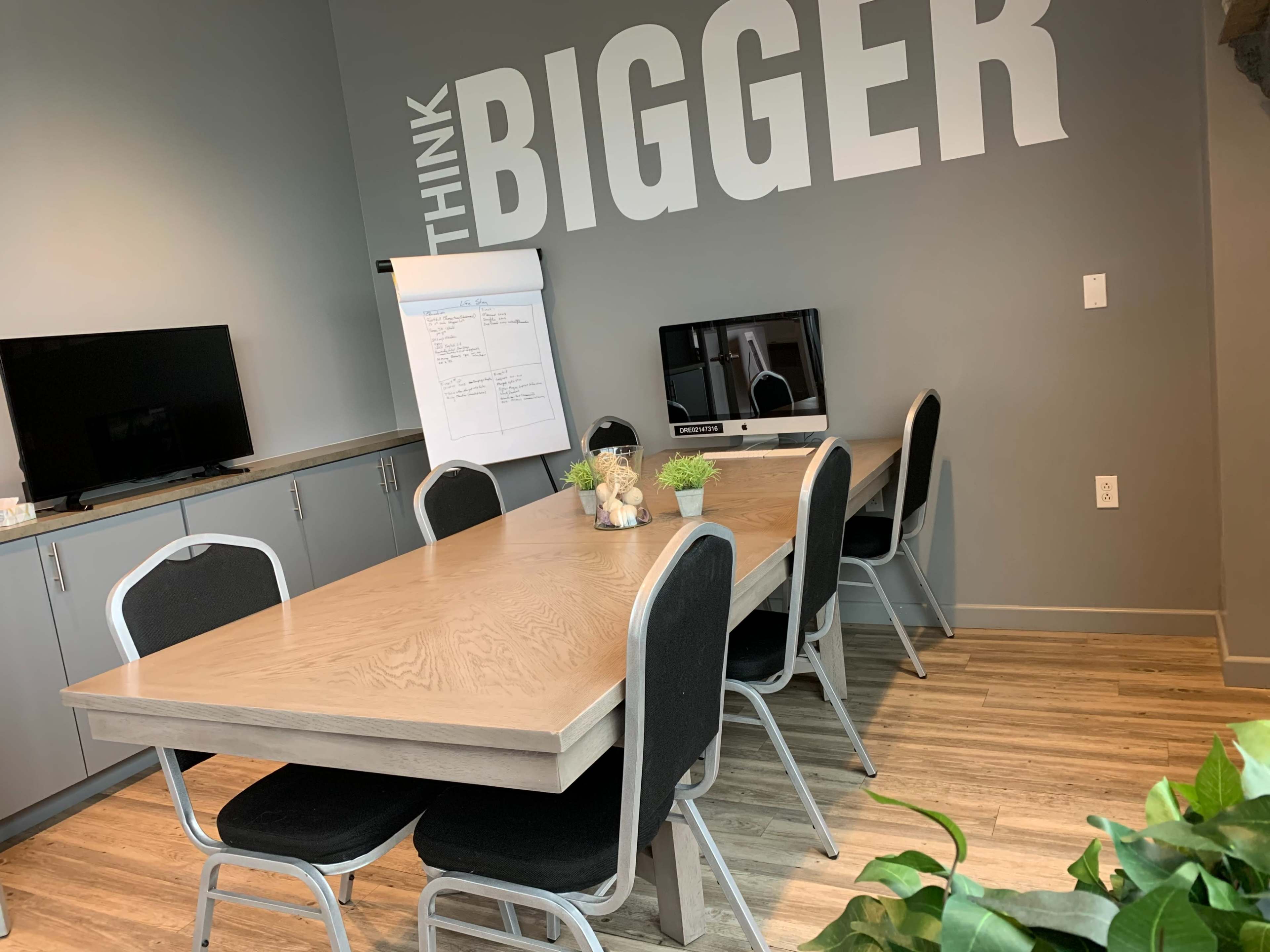 A conference room features a large wooden table surrounded by black chairs, with a whiteboard and two screens on the walls.