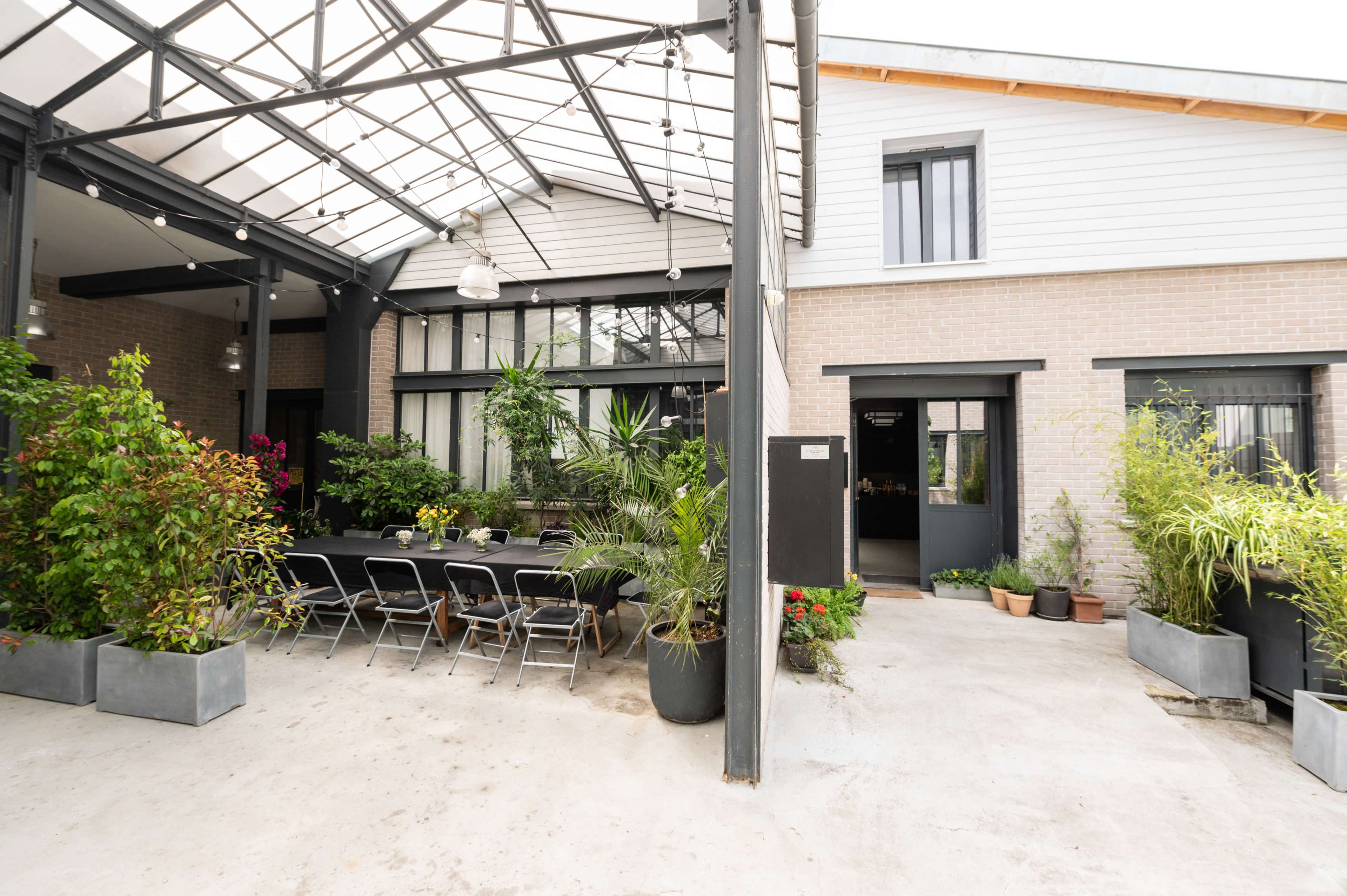 The image shows a modern outdoor dining area with a long table surrounded by greenery and potted plants, adjacent to a building with large windows and a metal roof structure.
