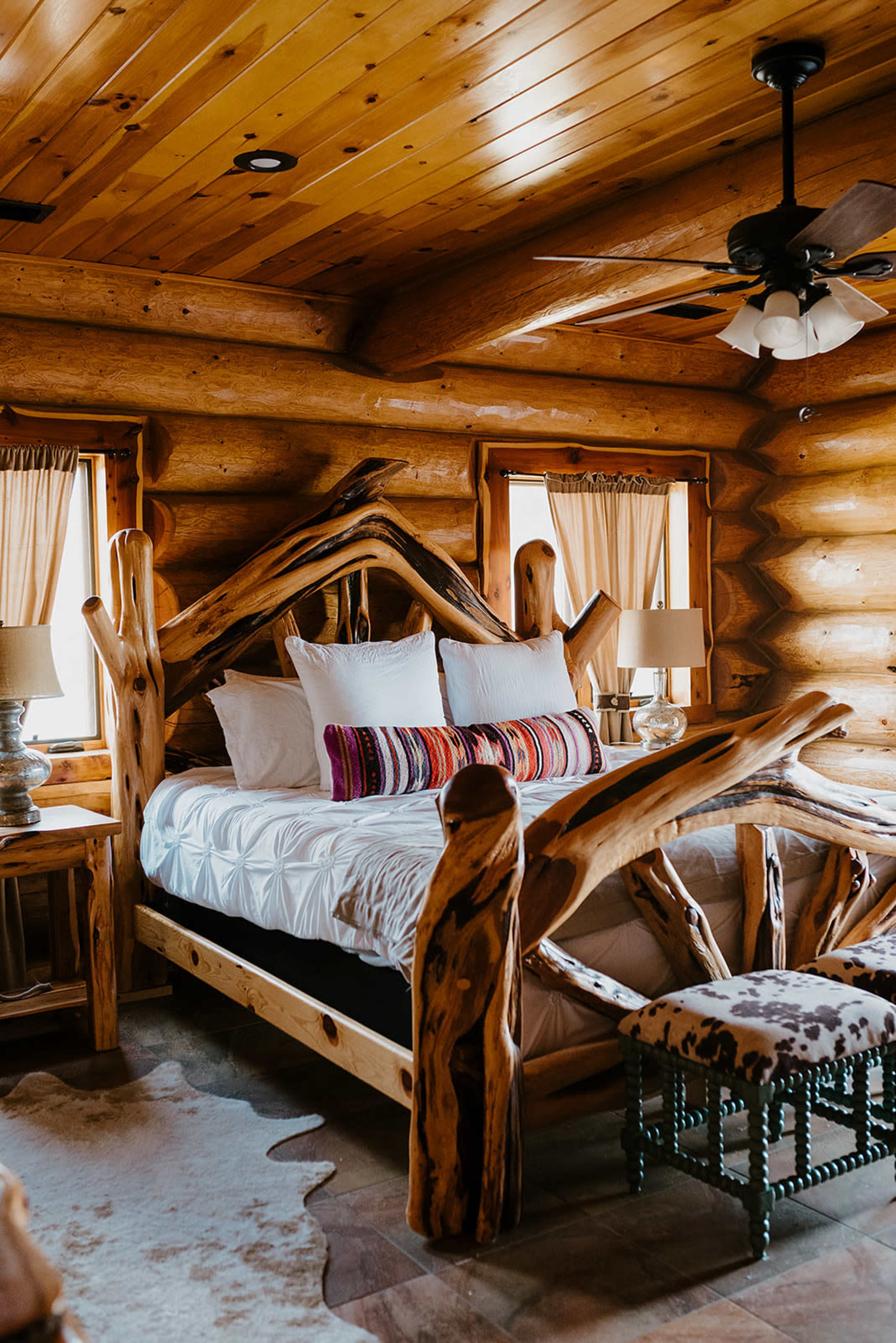 The image shows a rustic log cabin bedroom featuring a wooden bed frame with intricate branch-like details, a white bedspread, and two bedside lamps.