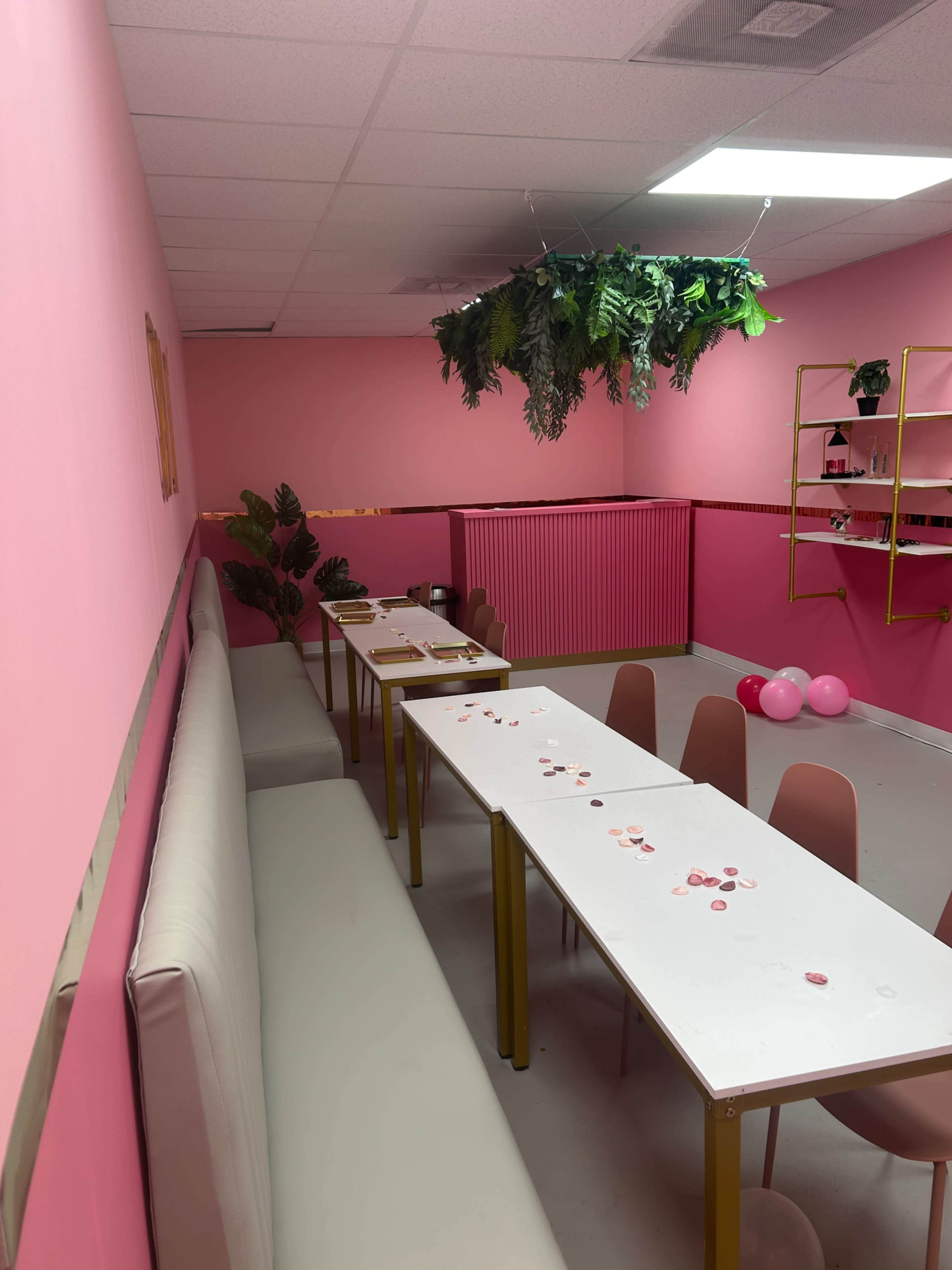 The image shows a pink-themed room with long white tables, chairs, and decorations, including a green foliage arrangement hanging from the ceiling.