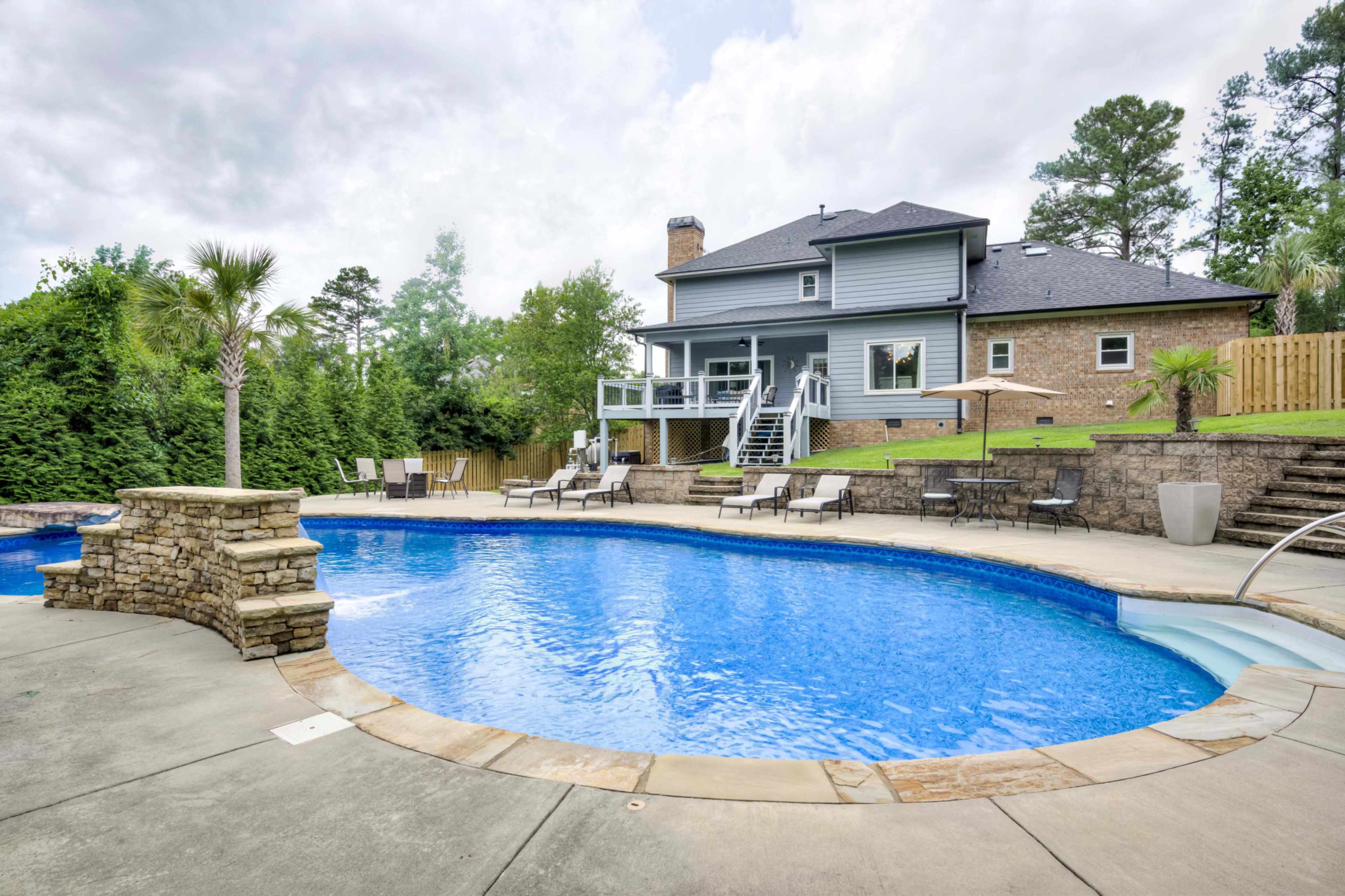 A large, rectangular swimming pool is surrounded by lounge chairs and features a stone wall, with a two-story house visible in the background amidst trees.