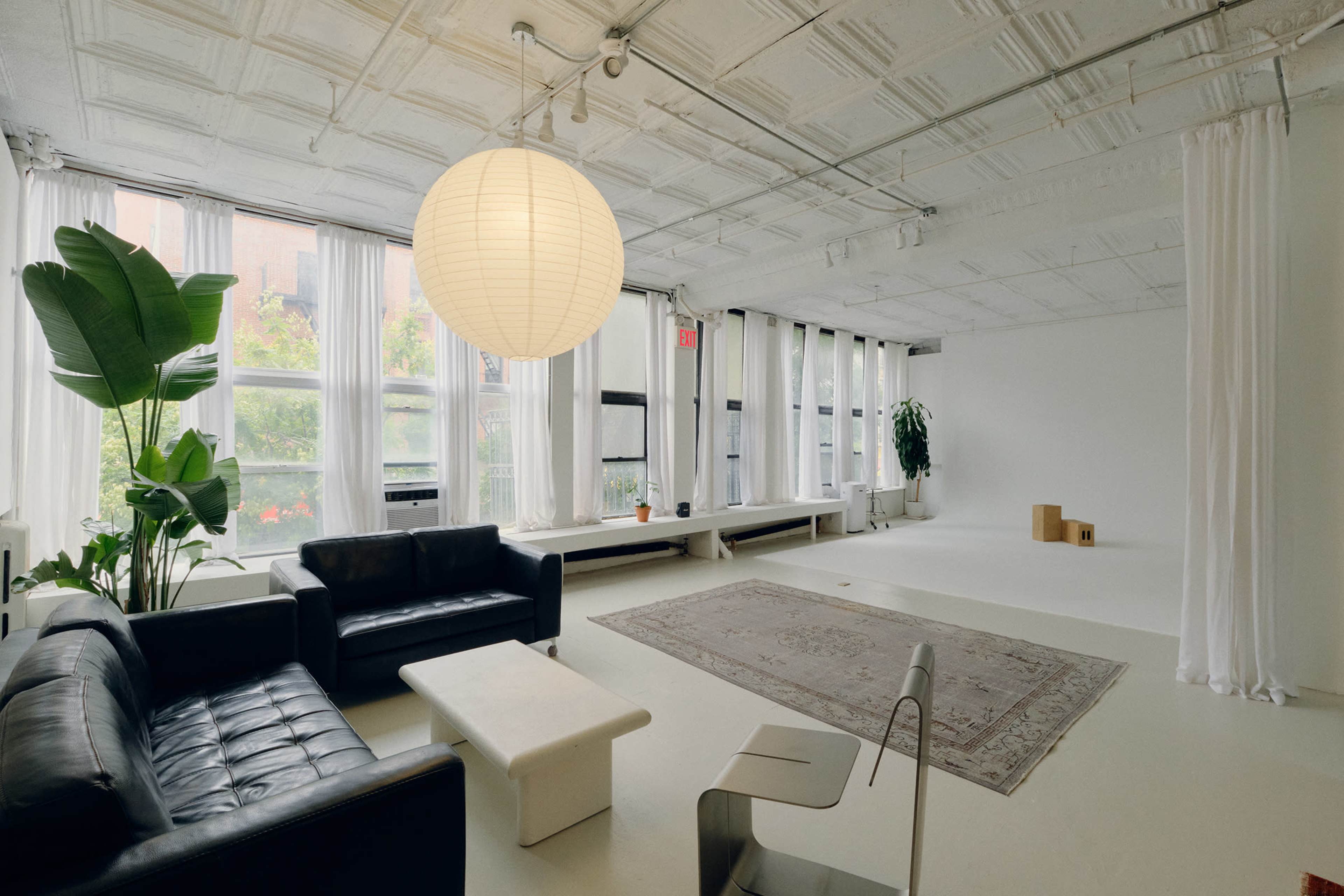 The image shows a spacious, minimalist living area featuring a black leather sofa, a white coffee table, and large windows with sheer curtains.