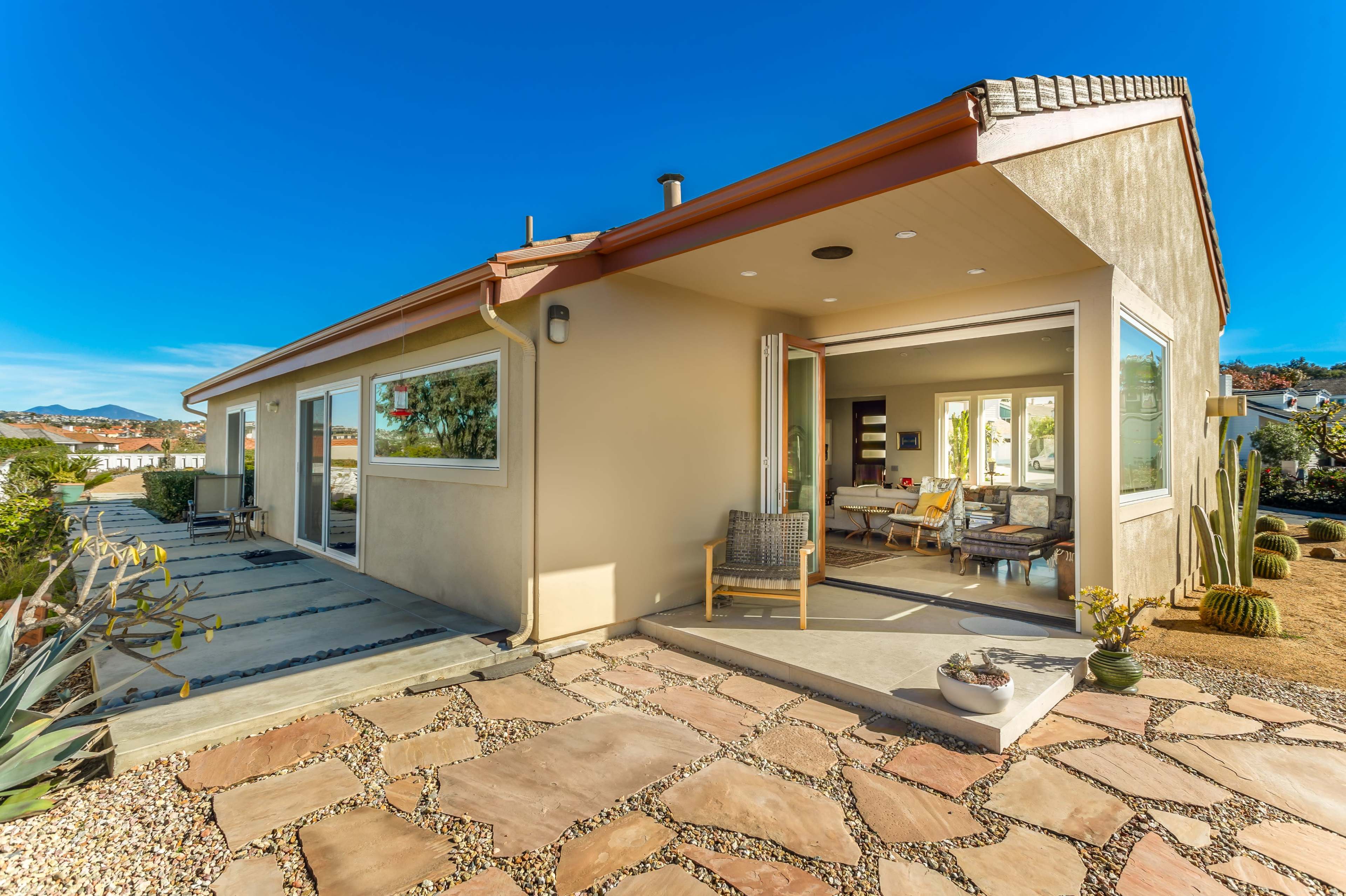 A modern single-story house features large windows, a patio with stone flooring, and a landscaped yard with cacti.