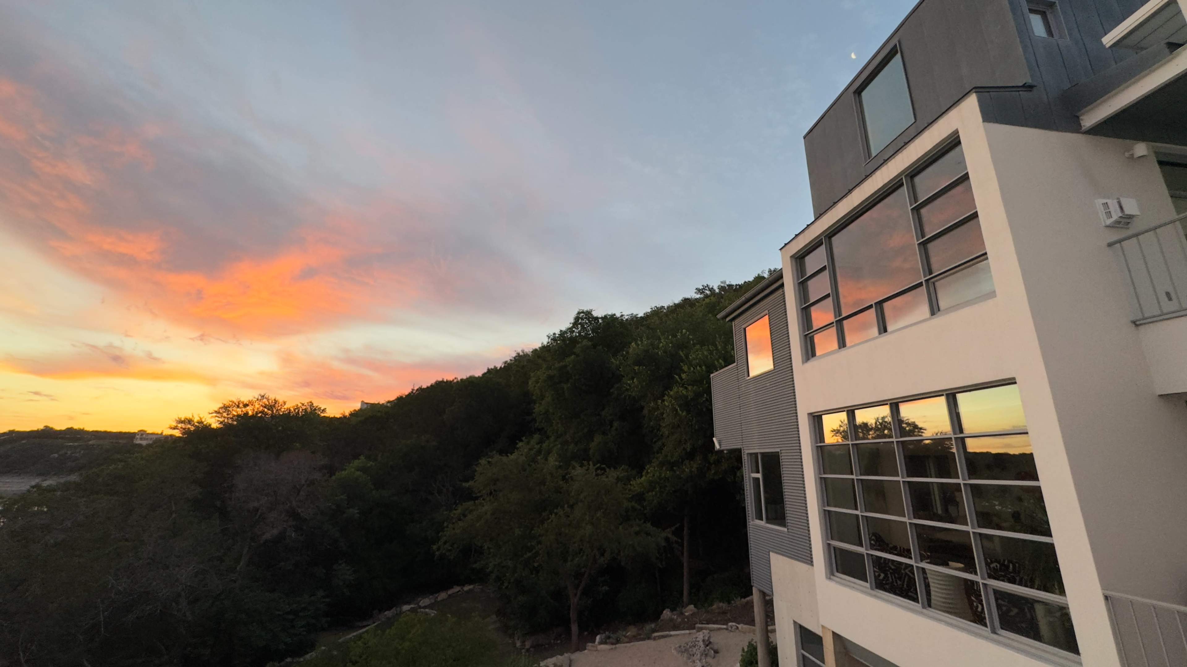 A modern house with large windows reflects a colorful sunset over a wooded area.