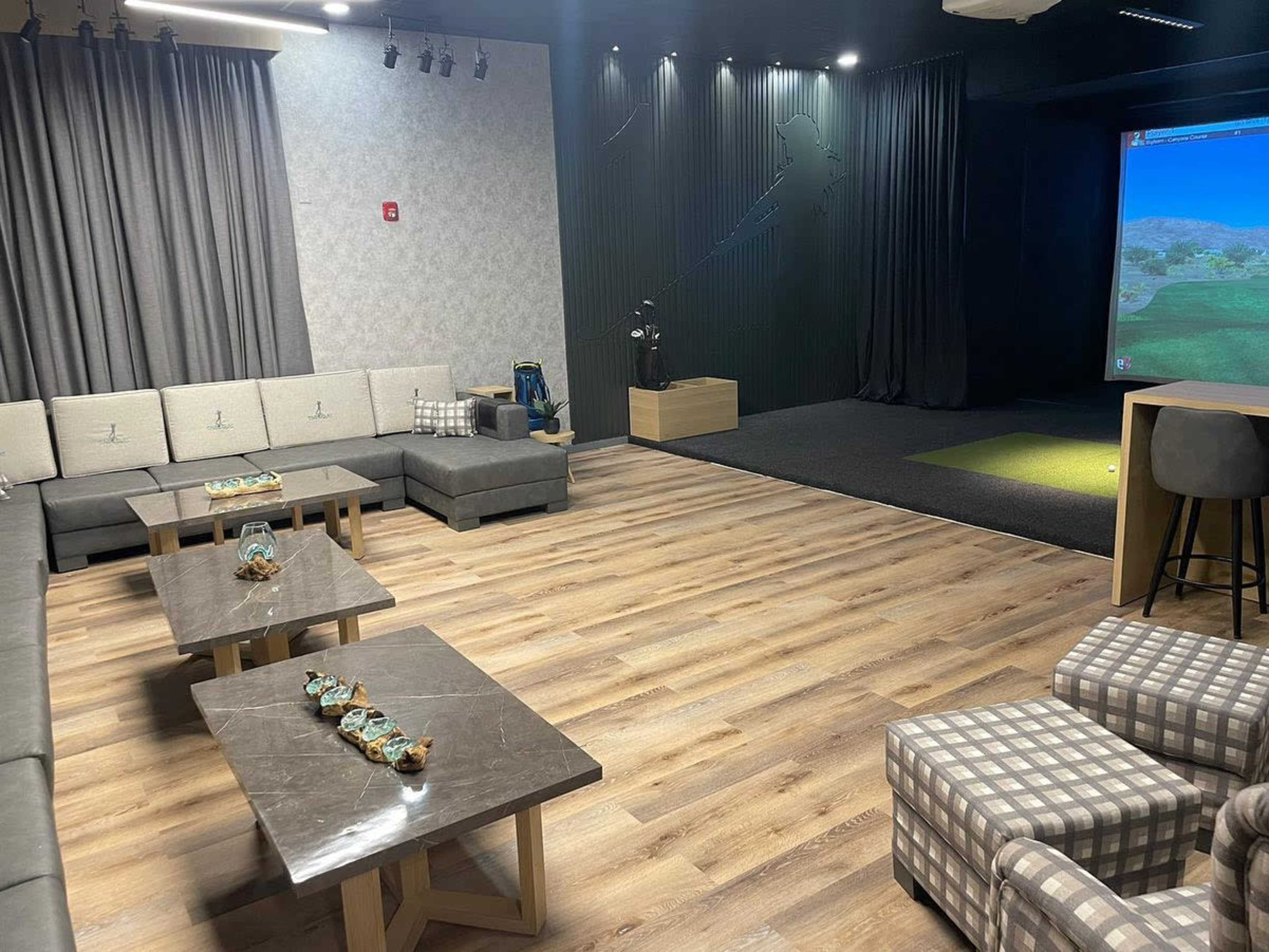 A spacious indoor lounge with seating arrangements featuring gray couches and wooden tables, alongside a golf simulation setup on one wall.