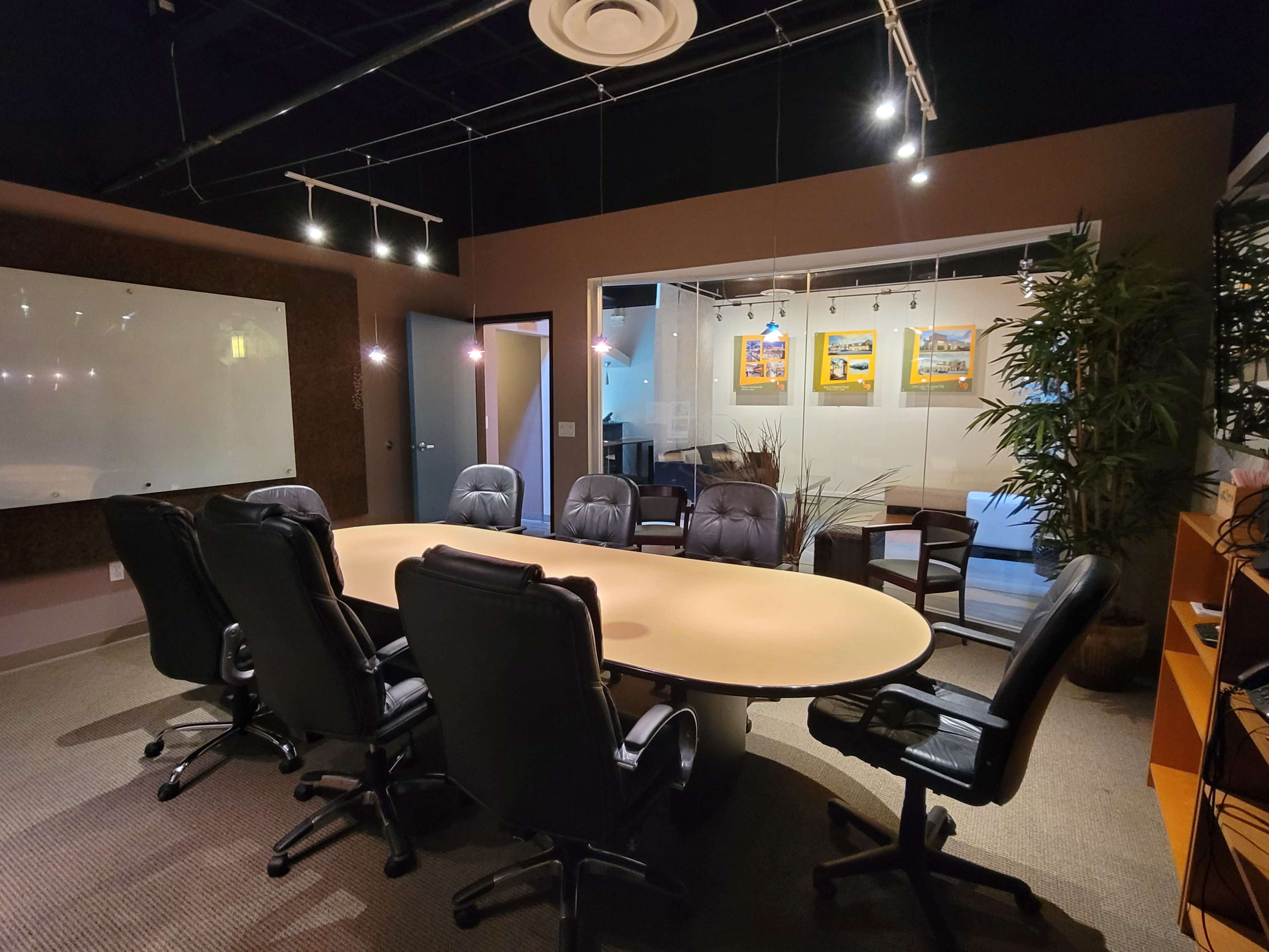 A conference room features a large oval table surrounded by black office chairs, with a glass wall showcasing artwork and a seating area in the background.