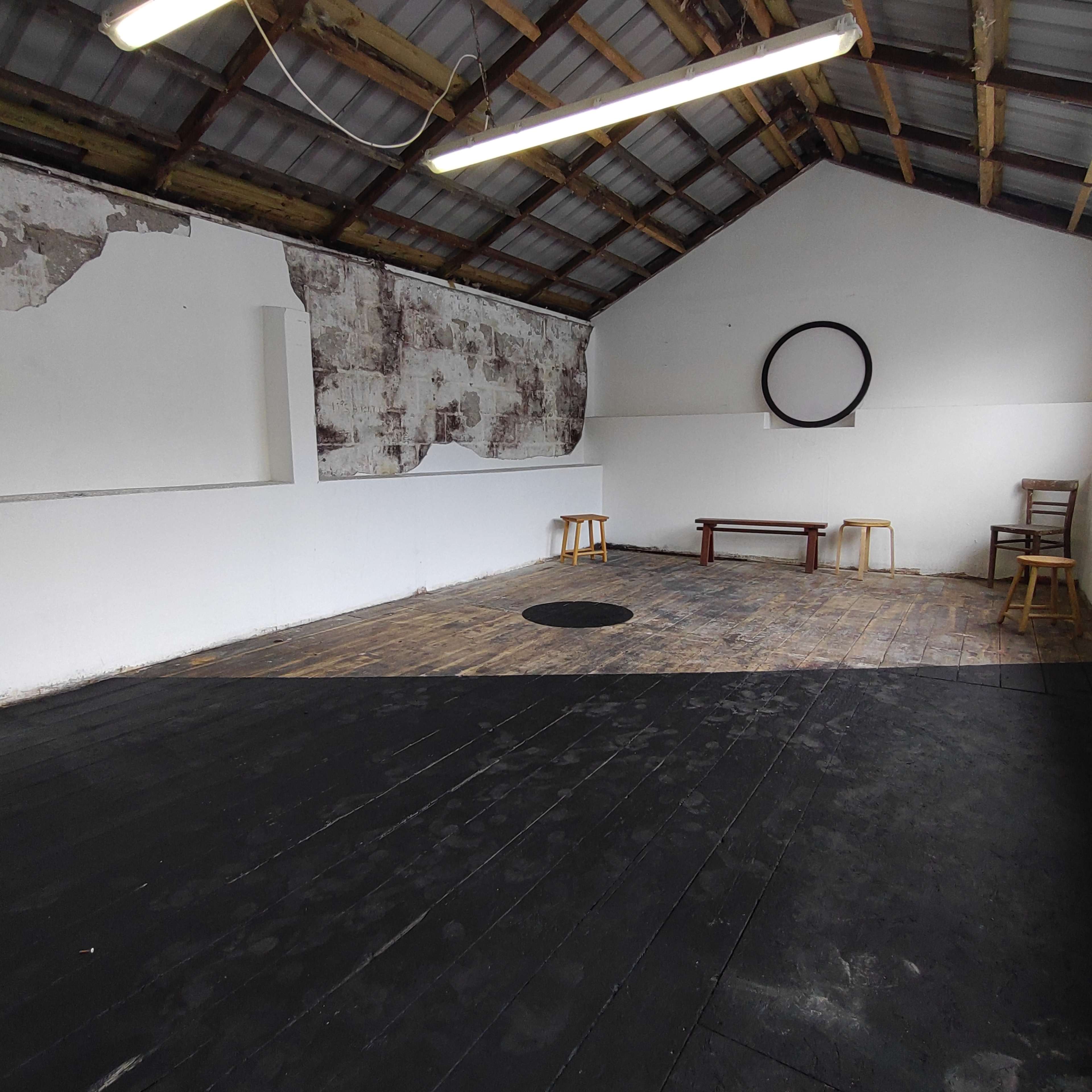 A spacious interior with wooden flooring, bare walls, and minimal furniture including two stools and a table.