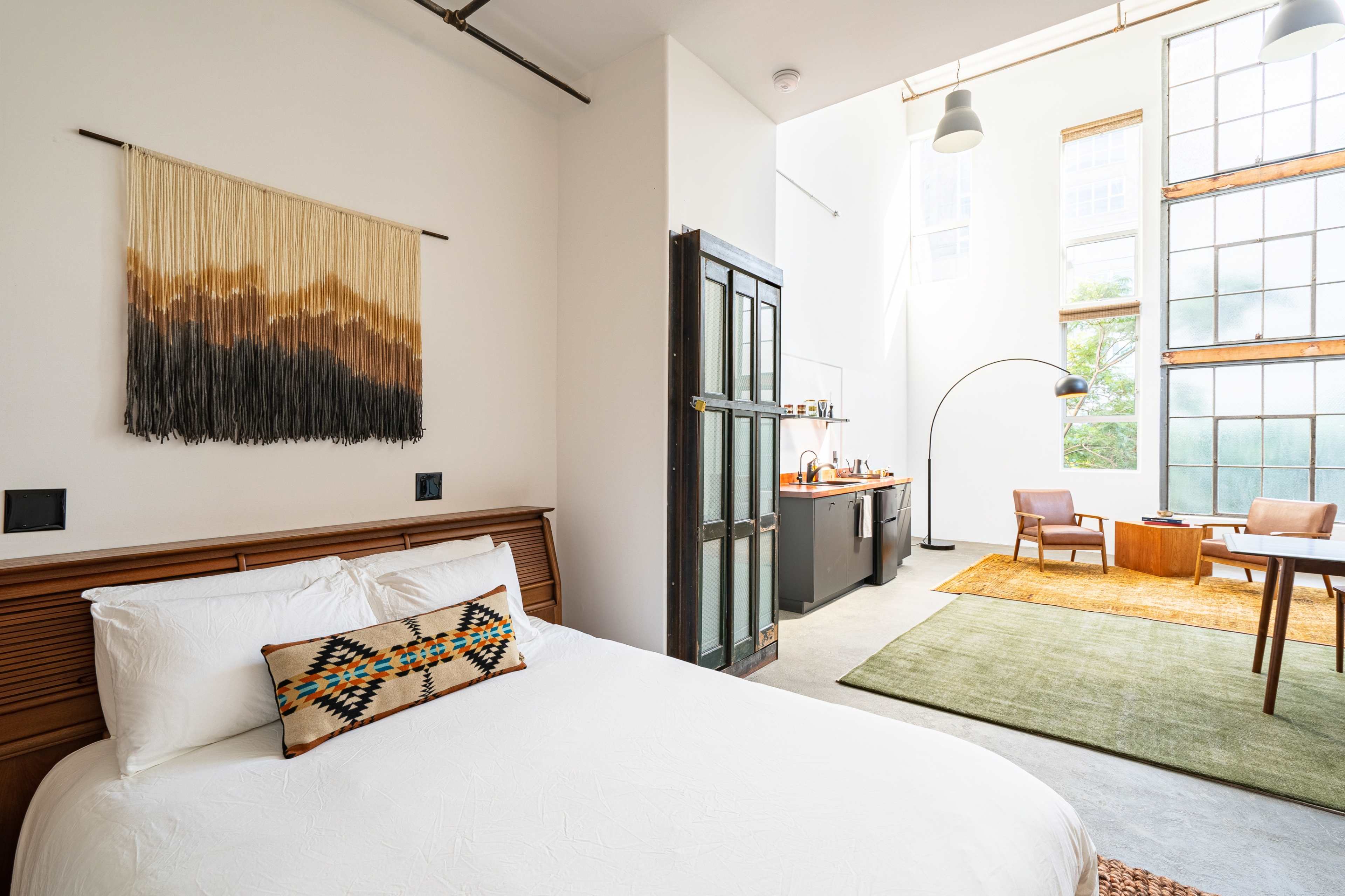 A bright, modern bedroom features a bed with decorative pillows, a hanging macramé wall piece, and connects to a spacious living area with large windows and minimalist furniture.