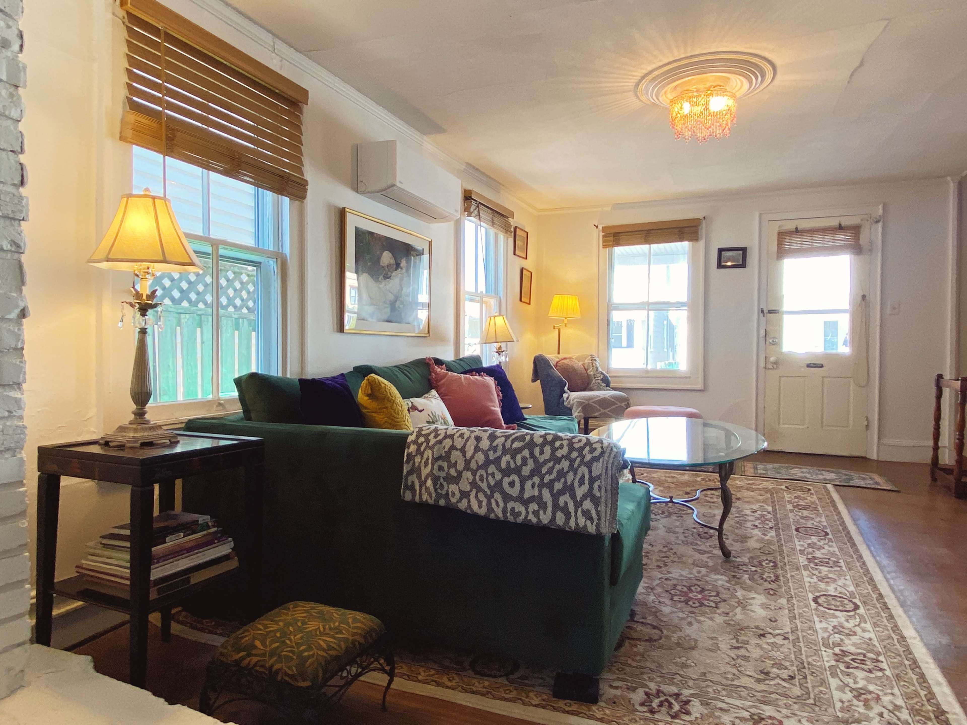 A living room with a green sofa, two armchairs, and a glass coffee table, illuminated by natural light from windows, with decorative lamps and artwork on the walls.