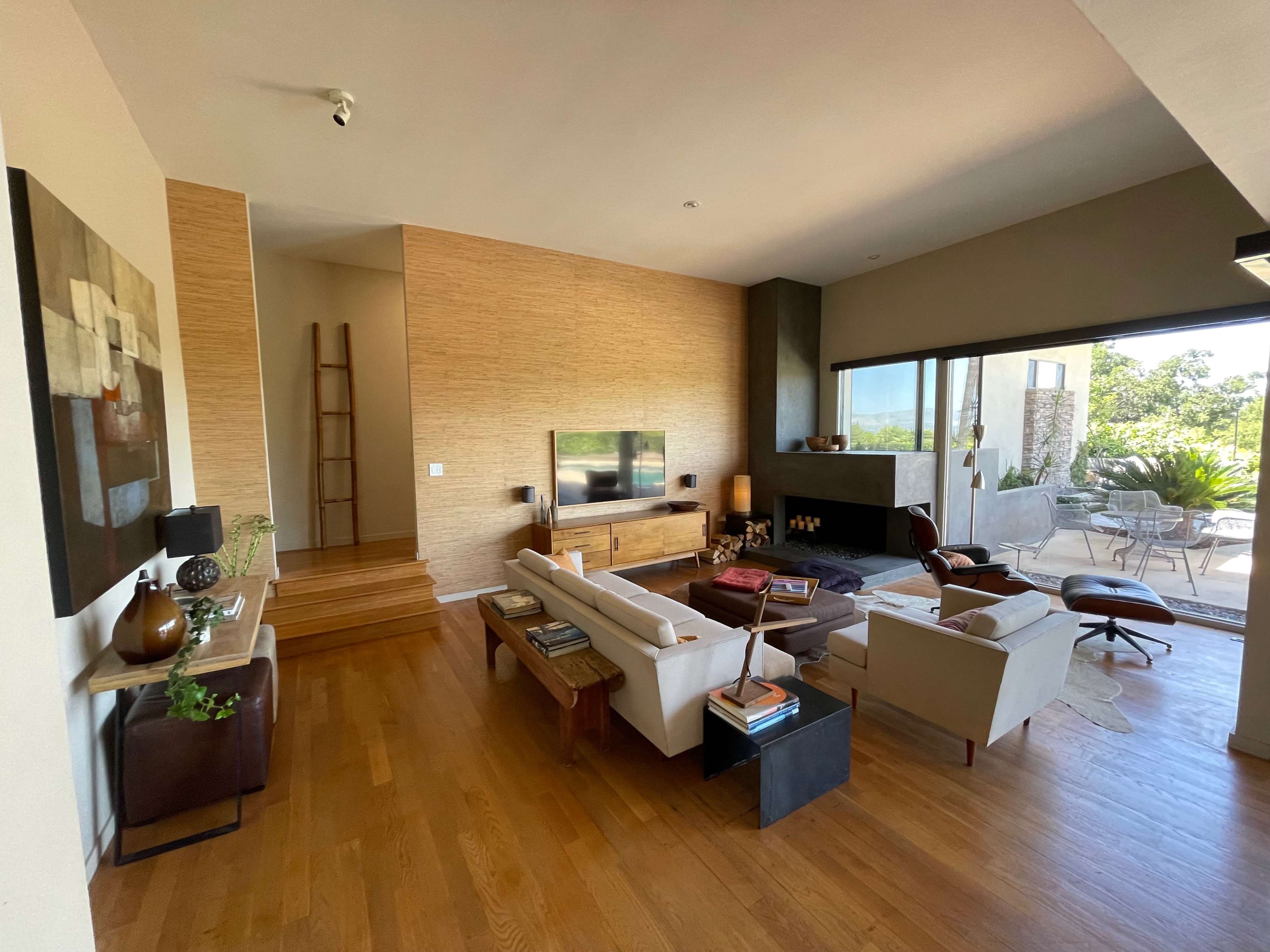 A modern living room with a large window, wooden flooring, and minimalistic furniture, leading to an outdoor patio.