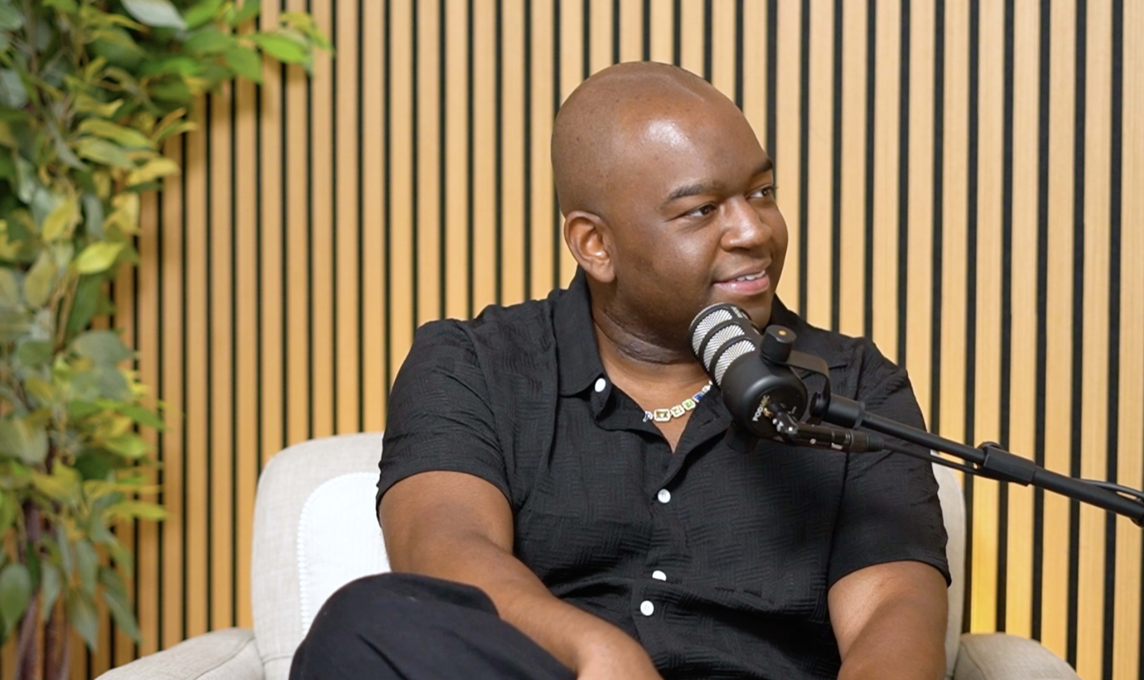 A man with a shaved head sits on a couch while speaking into a microphone in a modern setting with wooden paneling and plants in the background.