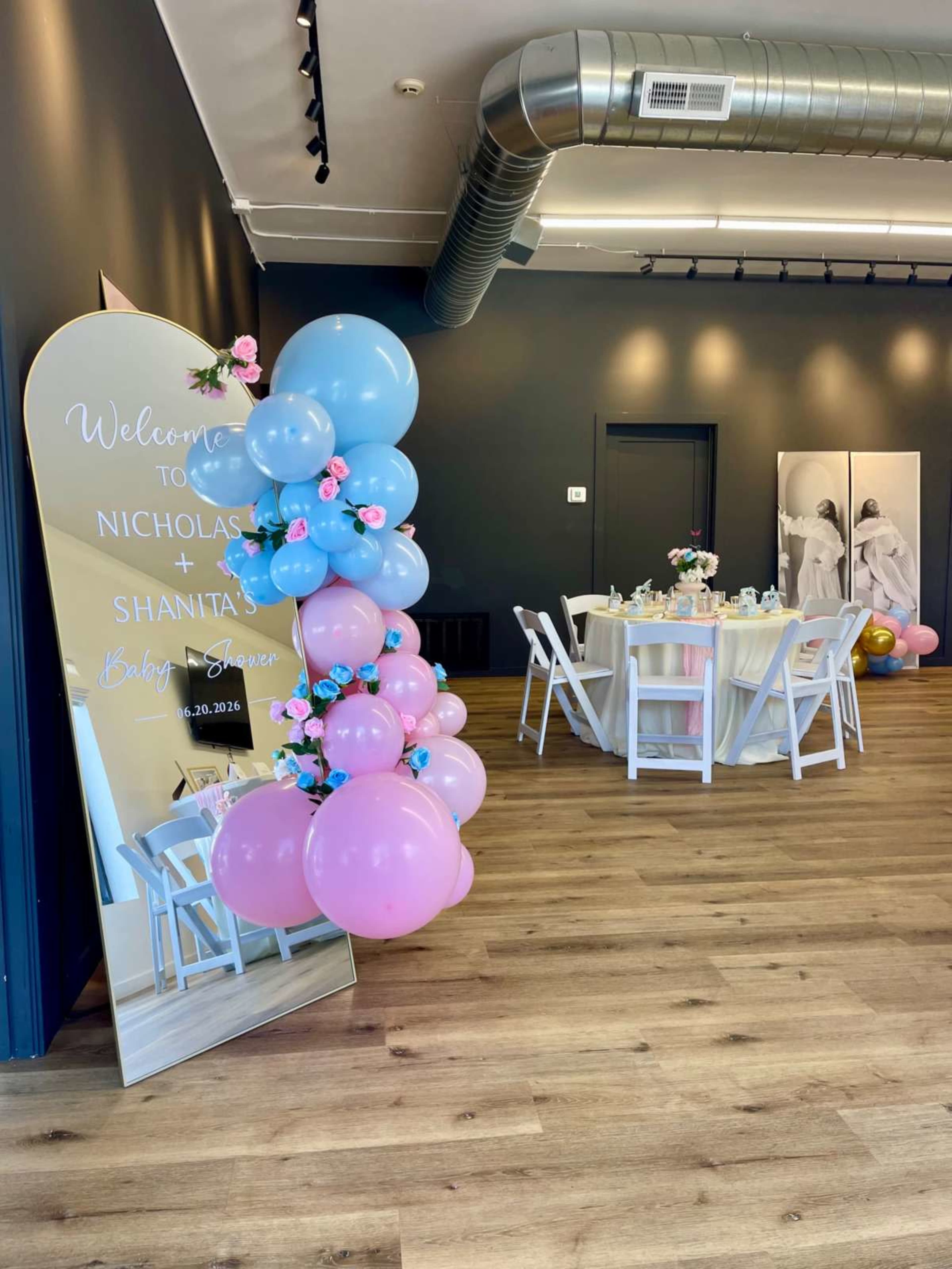 The image shows a decorated event space with a mirrored welcome sign and a balloon column in pink and blue next to a round dining table set for an occasion.