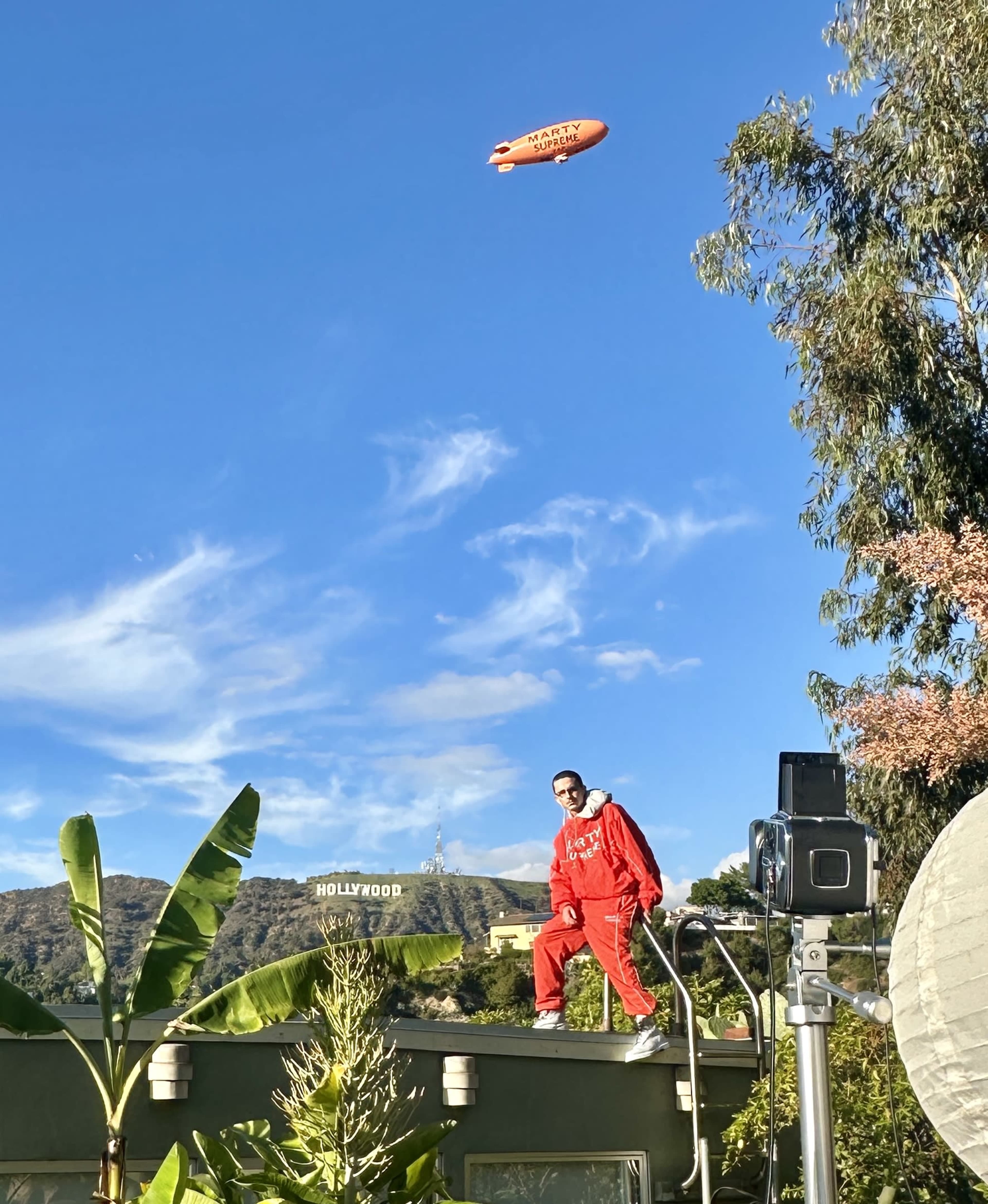 A person in a red jumpsuit stands on the roof of a building with the Hollywood sign visible in the background and a blimp flying overhead.