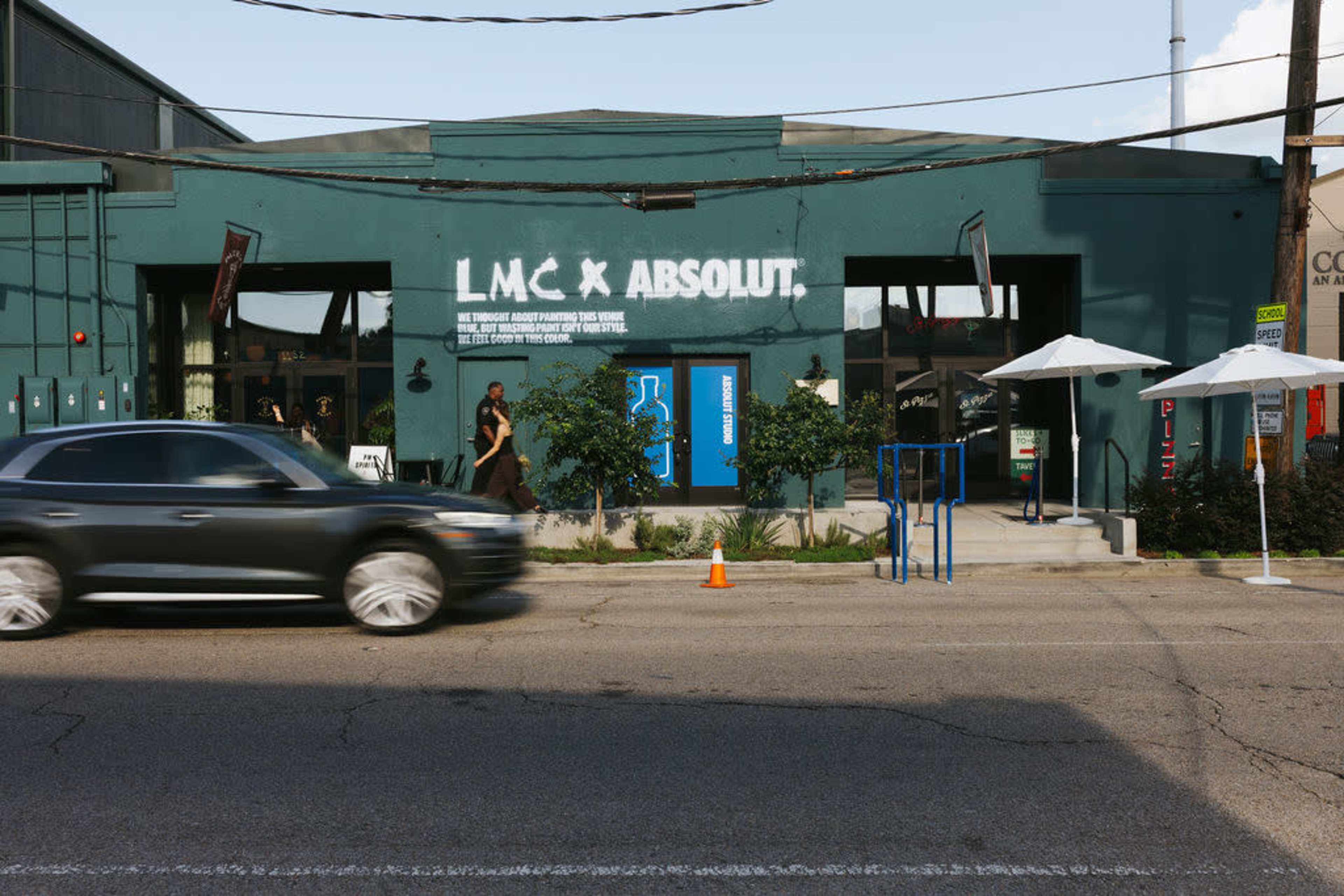 The image shows a teal-colored building with the text "L.M.C × ABSOLUT" displayed on its facade, while a black SUV drives past on the street.