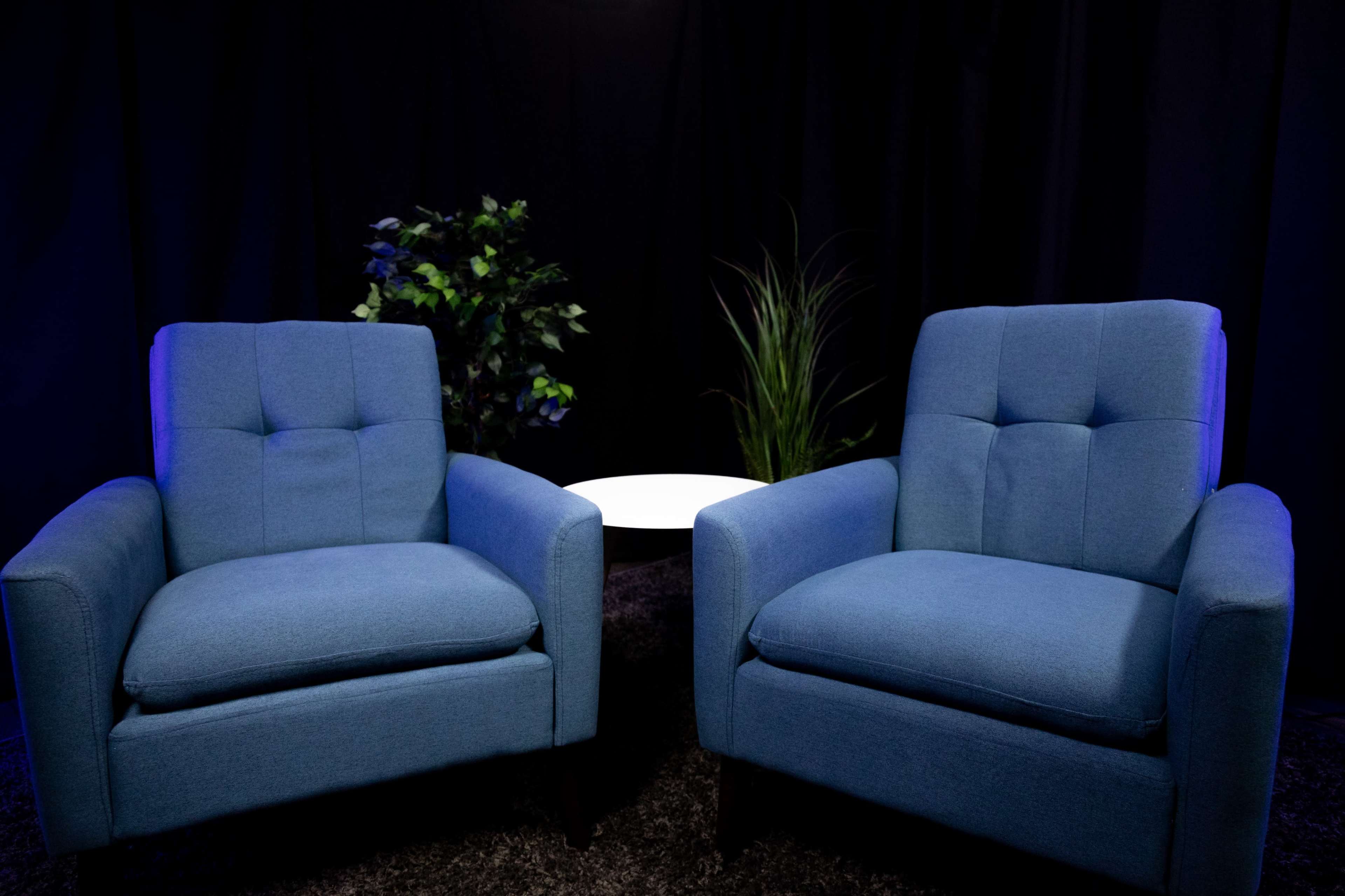 Two blue upholstered chairs are positioned facing each other beside a small white round table, with plants in the background.