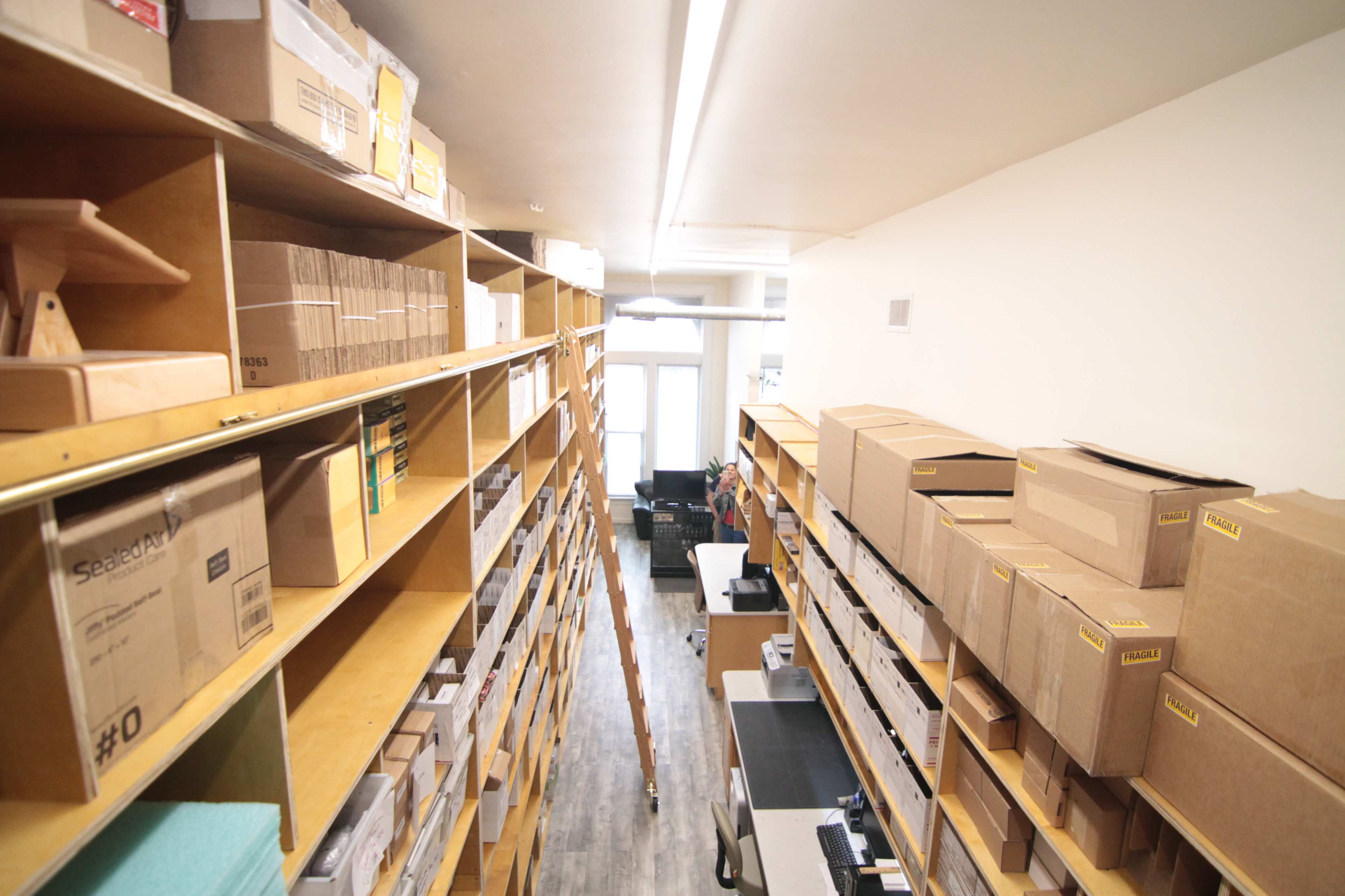 A narrow storage room features tall wooden shelves filled with boxes and packing supplies, leading to a workspace area at the far end.
