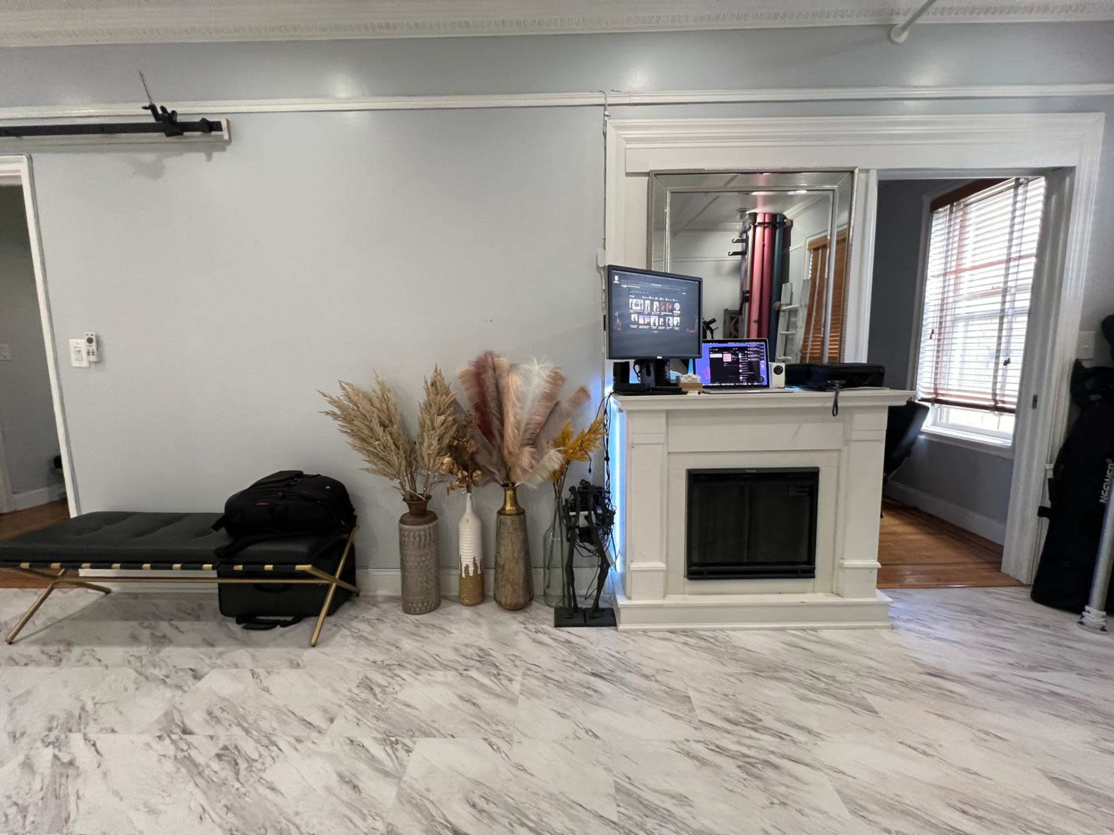 The image shows a modern interior with a sleek black bench, decorative dried plants in vases, a compact fireplace, and a desk area with a computer setup visible through a mirror.