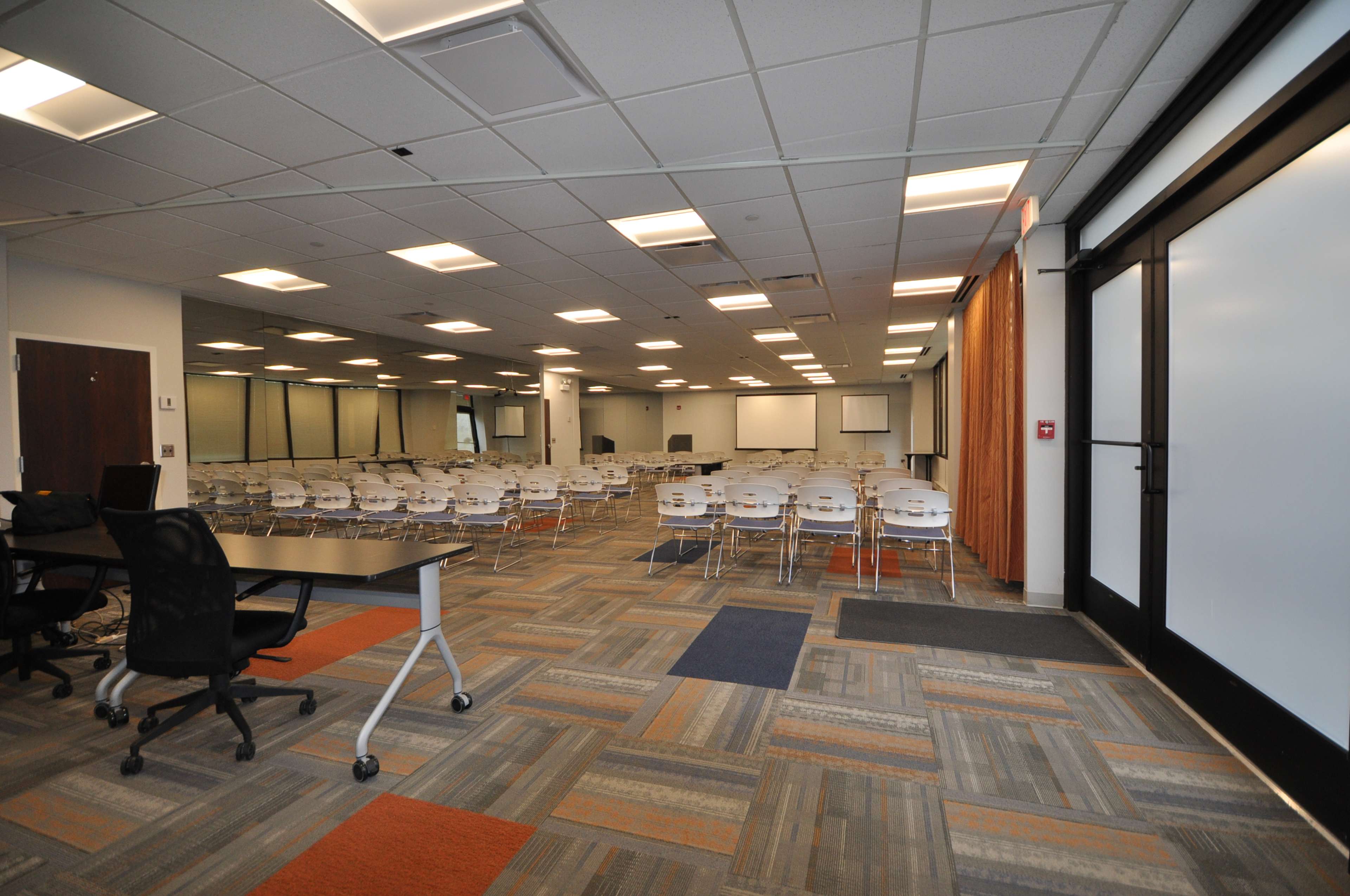 The image shows a spacious conference room set up with rows of white chairs facing a presentation area, with gray carpets and modern lighting.