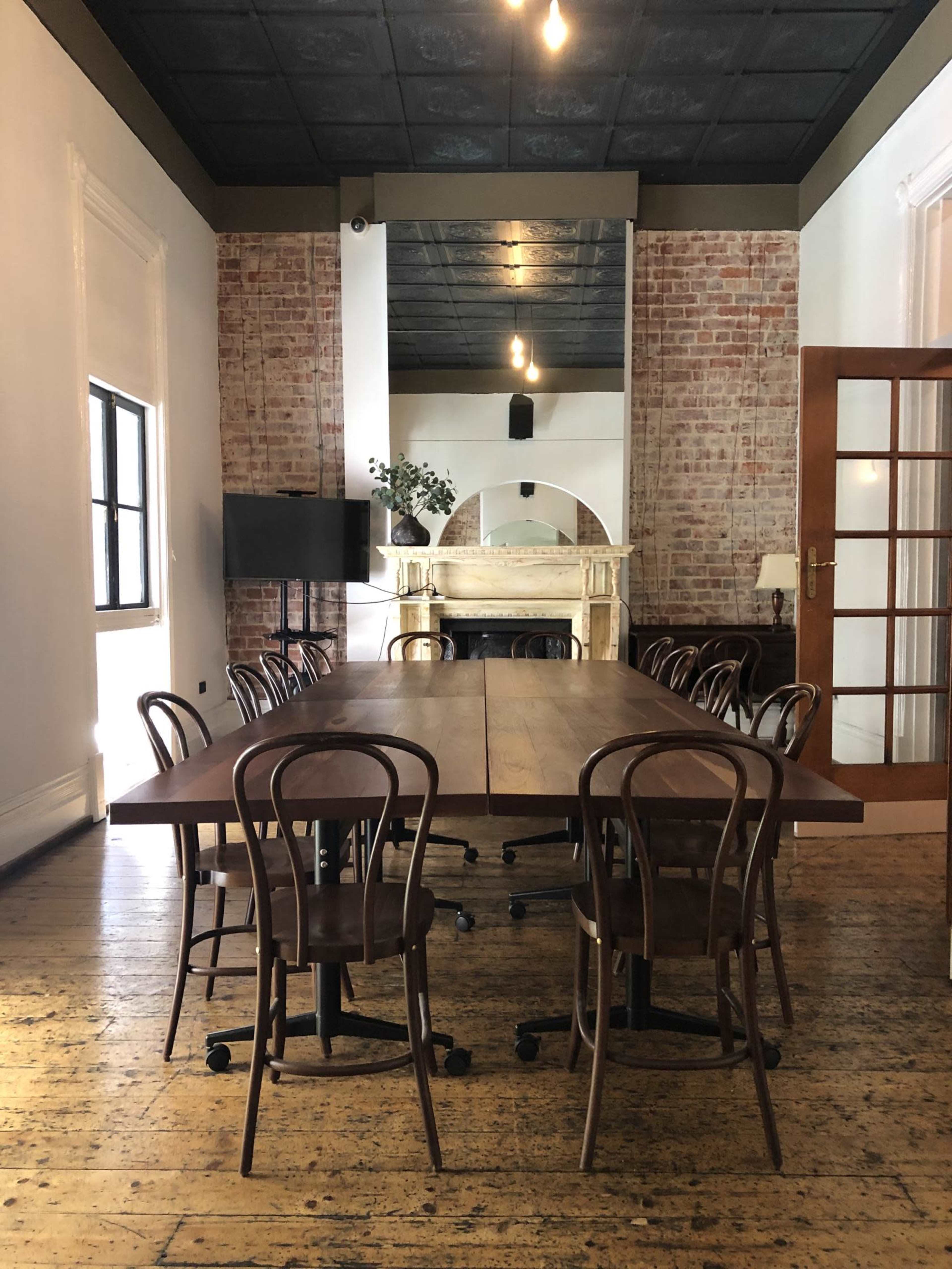 A long wooden table surrounded by rolling chairs is positioned in a spacious room with brick walls and large windows.