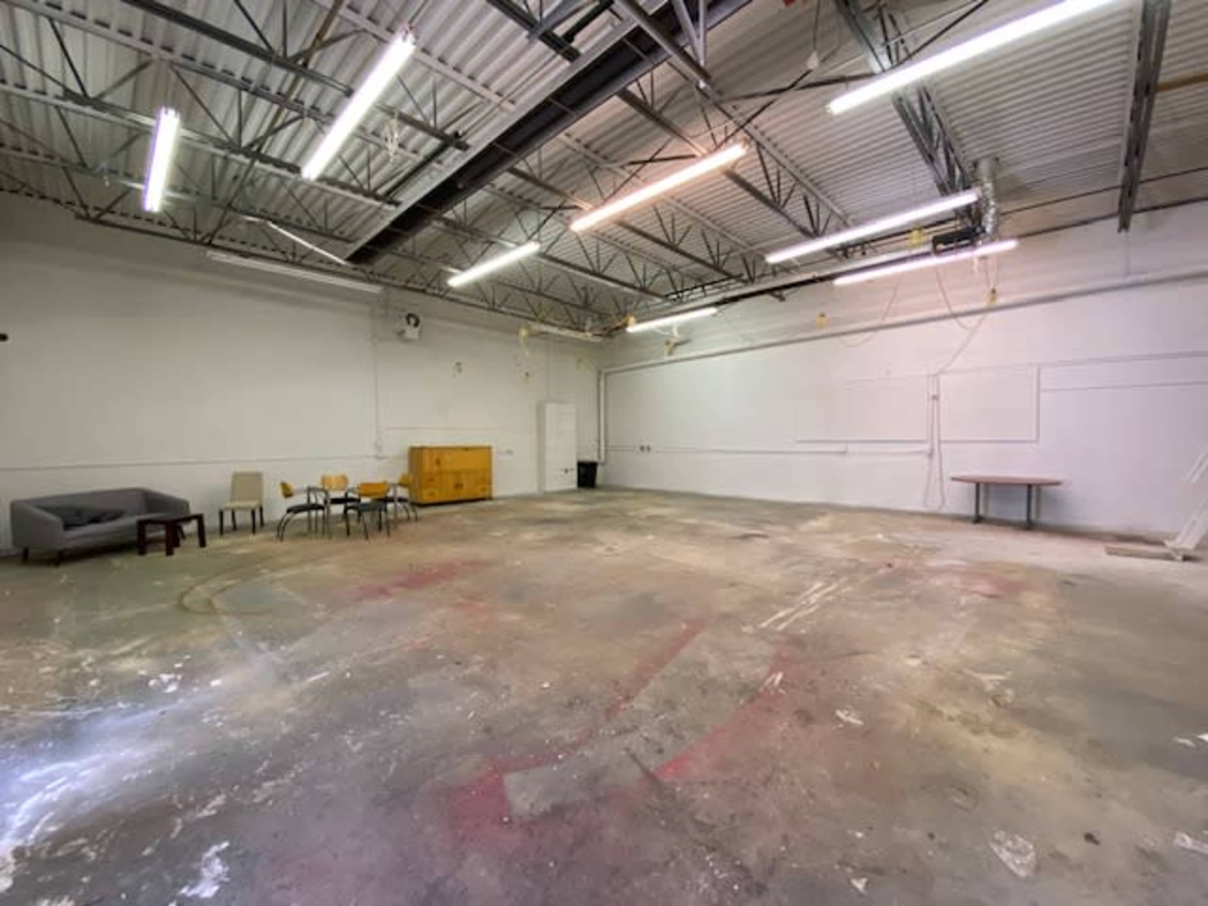 An empty industrial space features a concrete floor, scattered furniture, and overhead lighting.