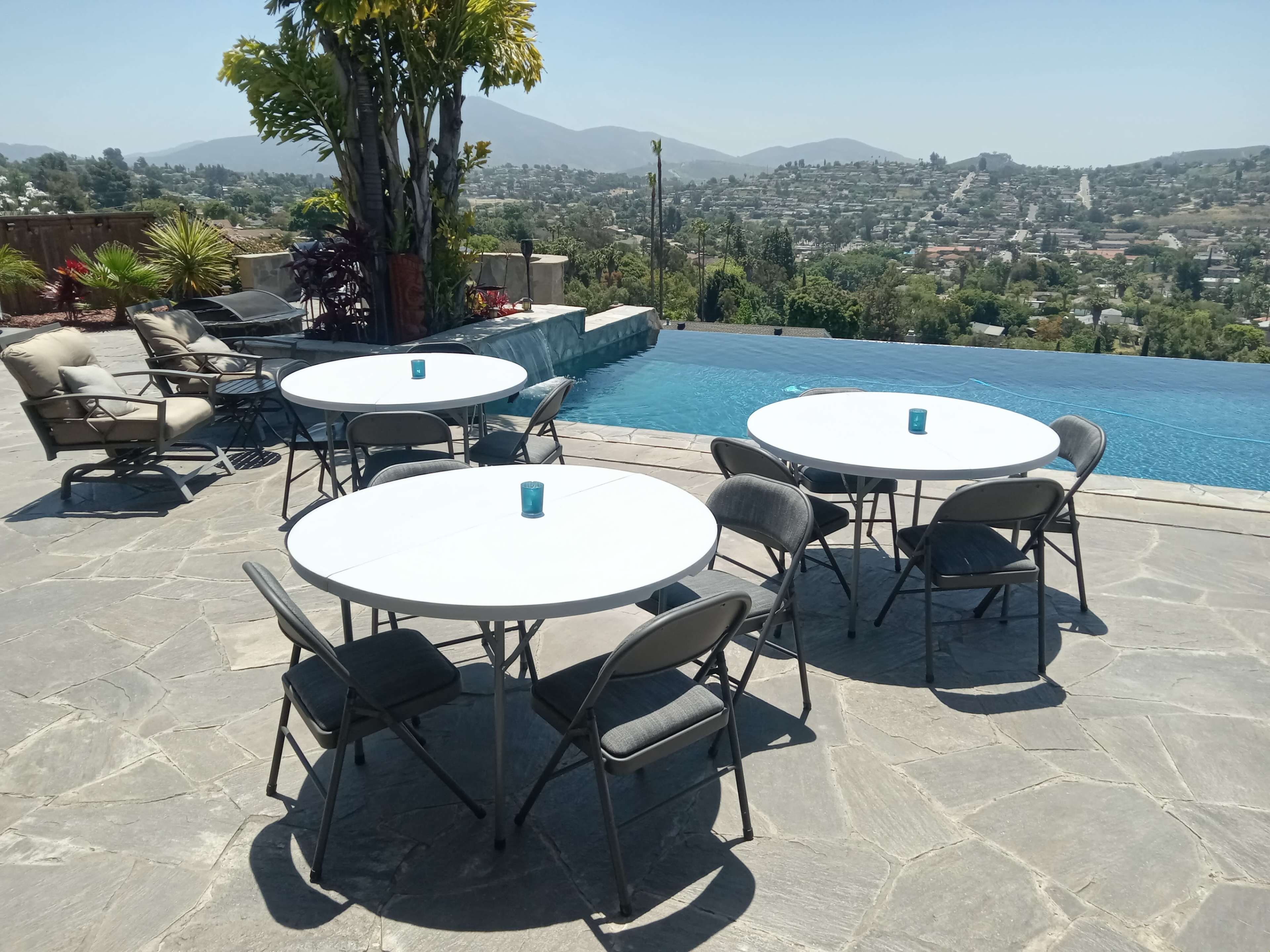 An outdoor patio area with several round tables and chairs overlooking a hillside view and an infinity pool.