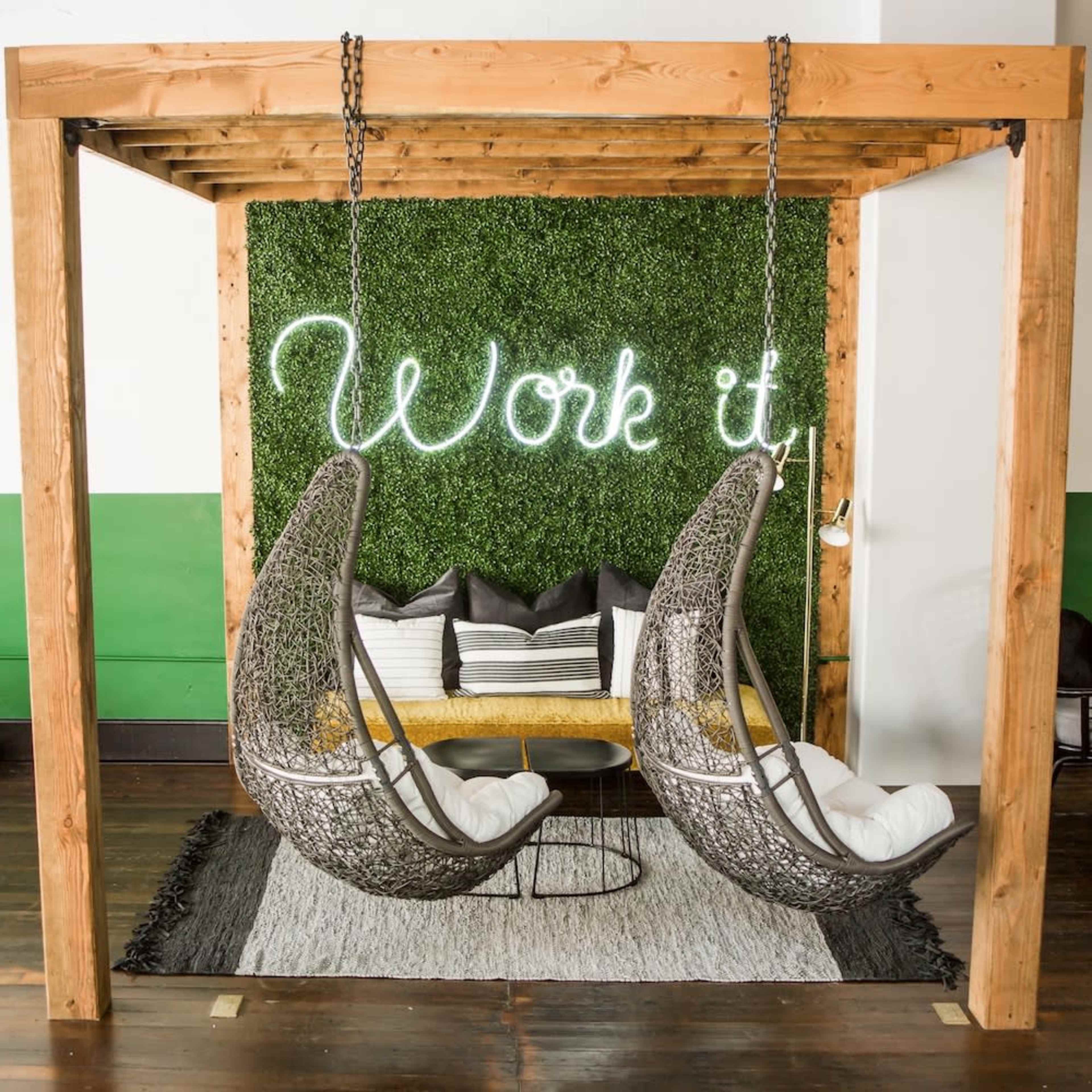 The image shows a wooden structure with two hanging chairs in front of a green wall adorned with a glowing neon sign that reads "Work it."