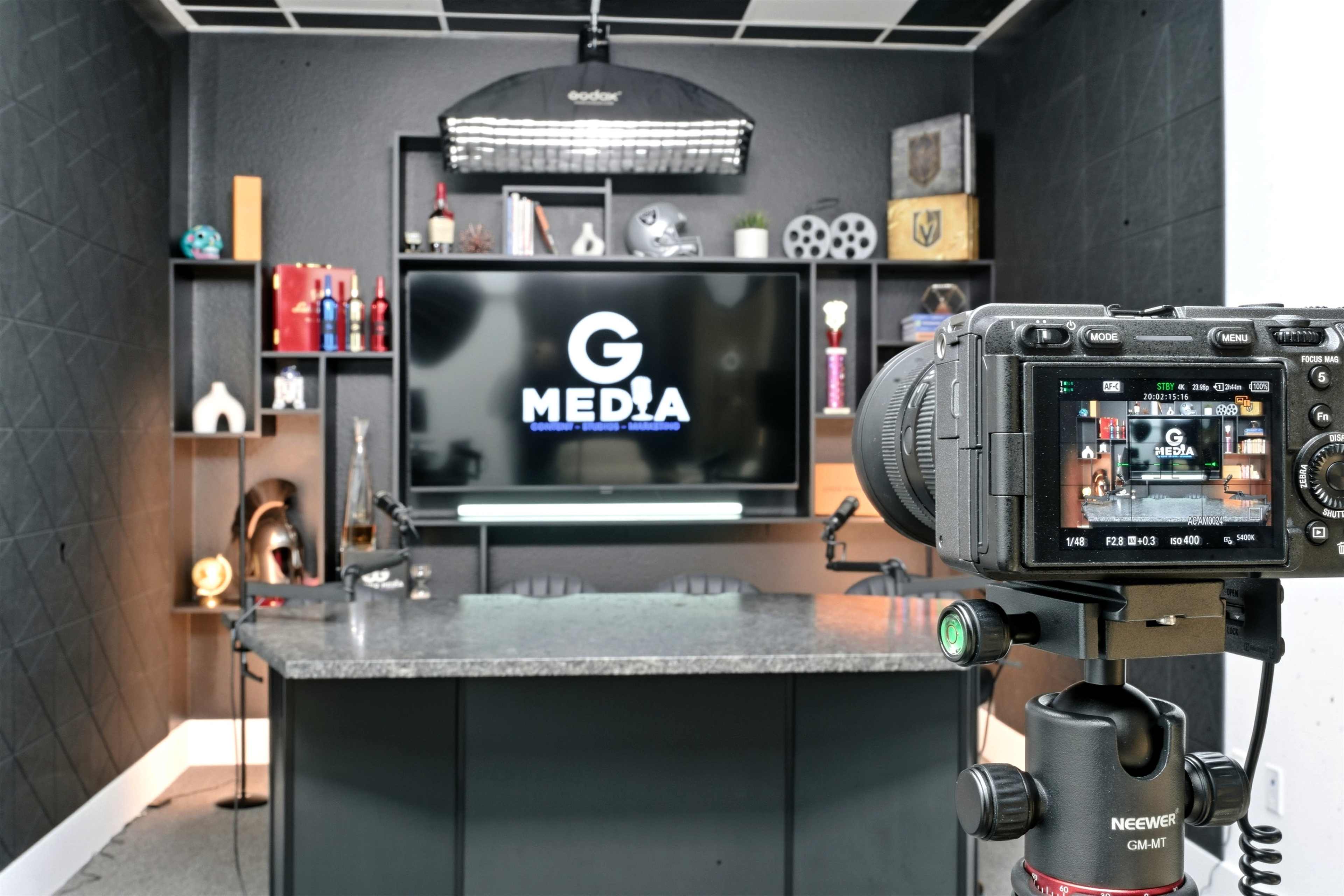 The image shows a media studio setup with a camera focused on a monitor displaying the logo "G MEDIA," surrounded by various decorative items on shelves.
