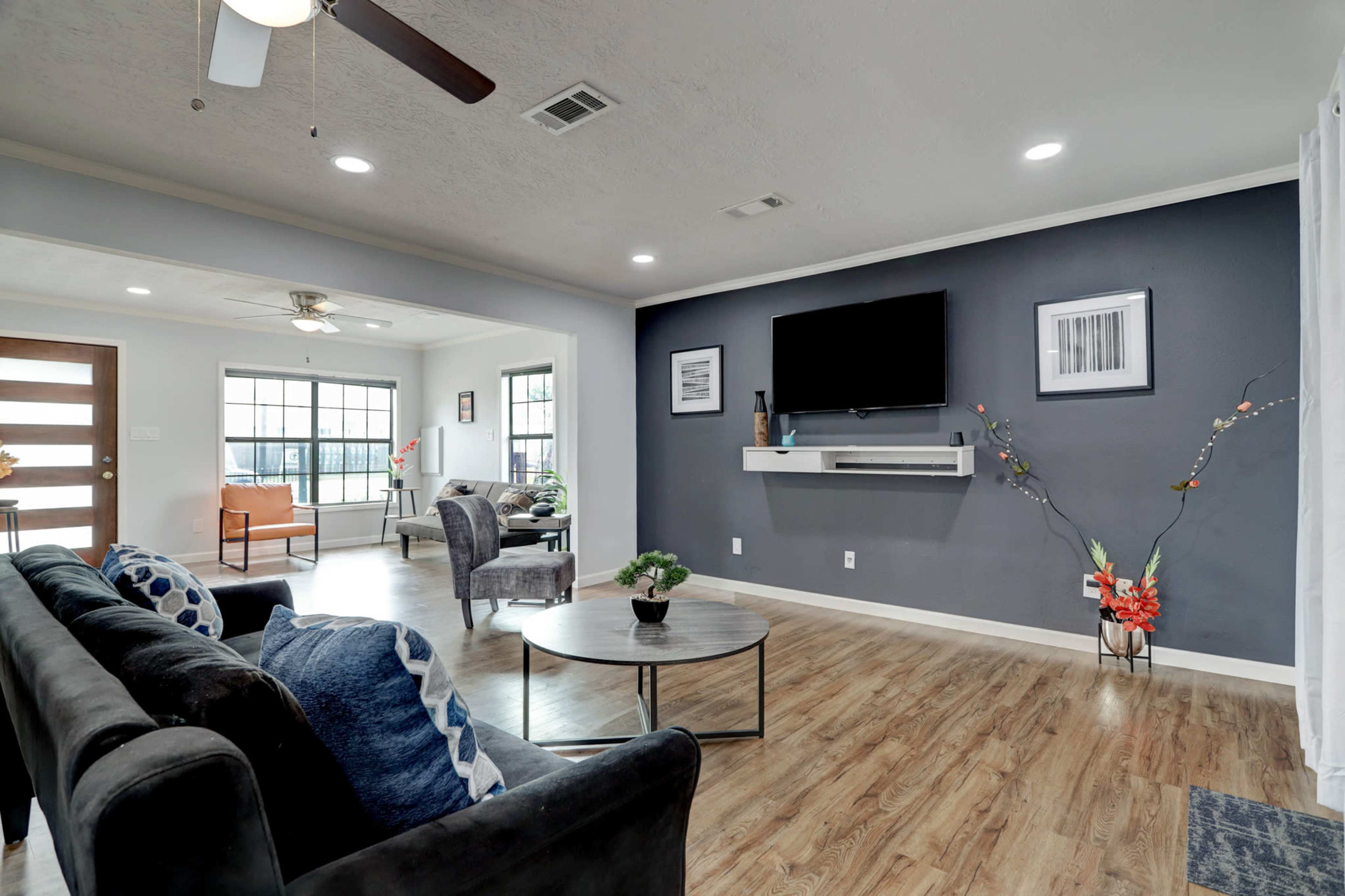A living room featuring a dark grey accent wall, a wall-mounted television, a coffee table, and seating arrangements including a black sofa and armchairs.