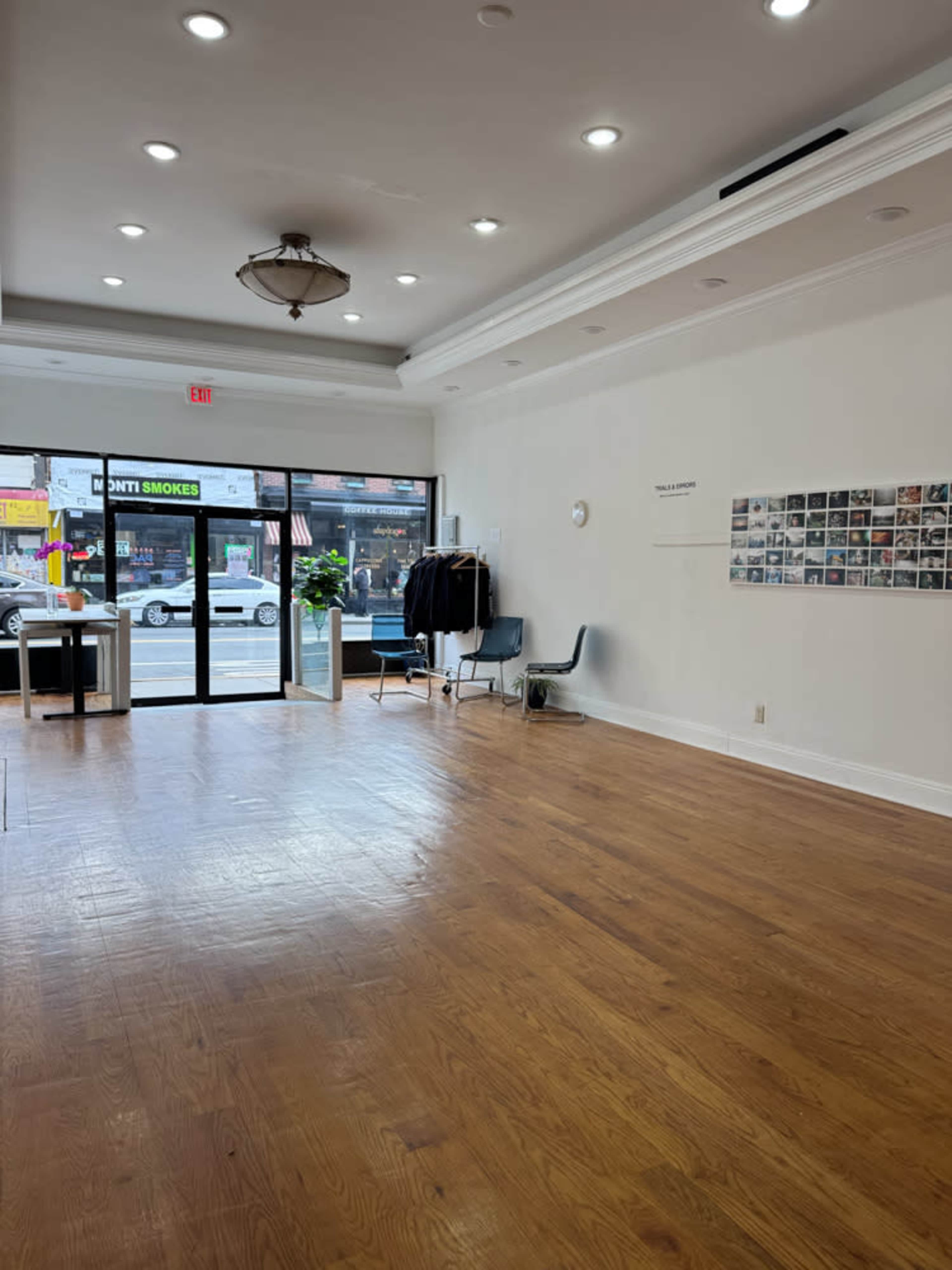 The image shows a spacious, brightly lit interior with wooden flooring, showcasing an empty room featuring a few chairs, a rack with coats, and a wall display of small photographs.