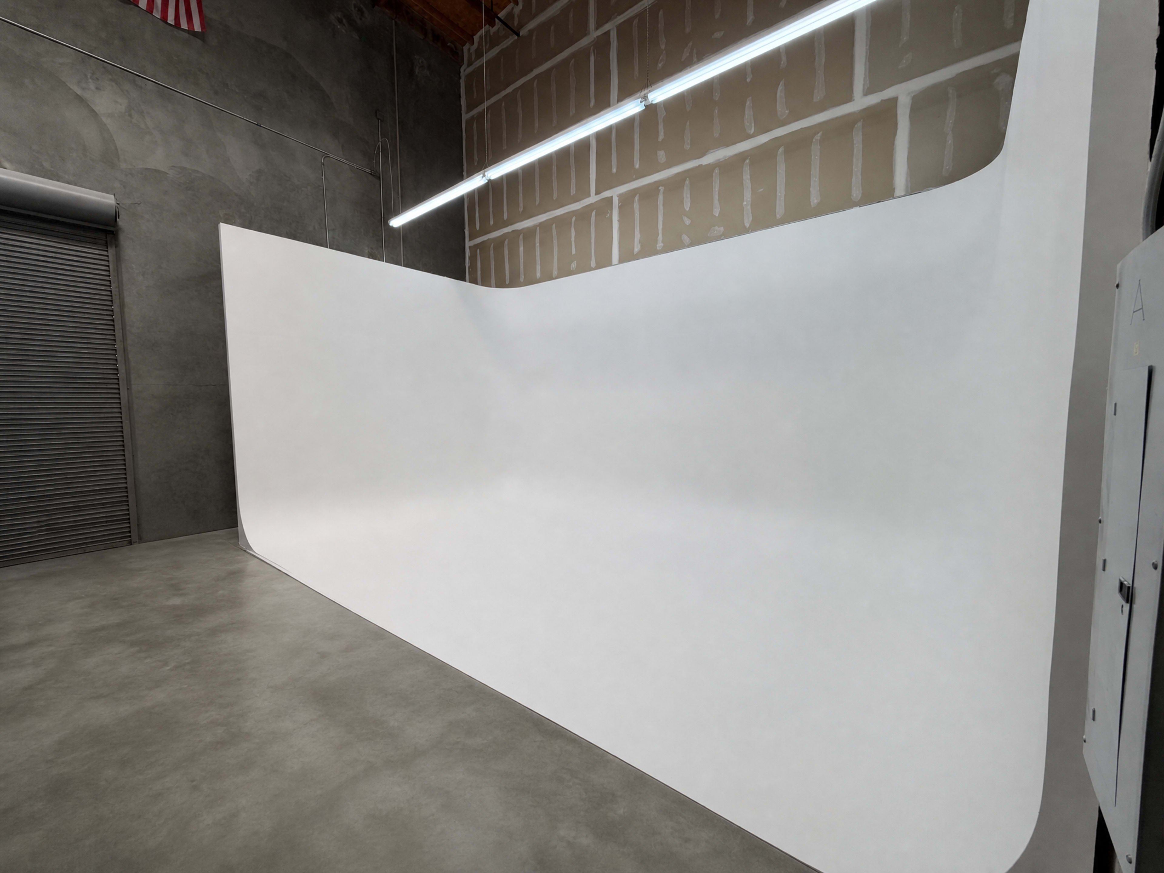 The image shows a large, curved white wall in a spacious room with concrete flooring and a metal roll-up door in the background.
