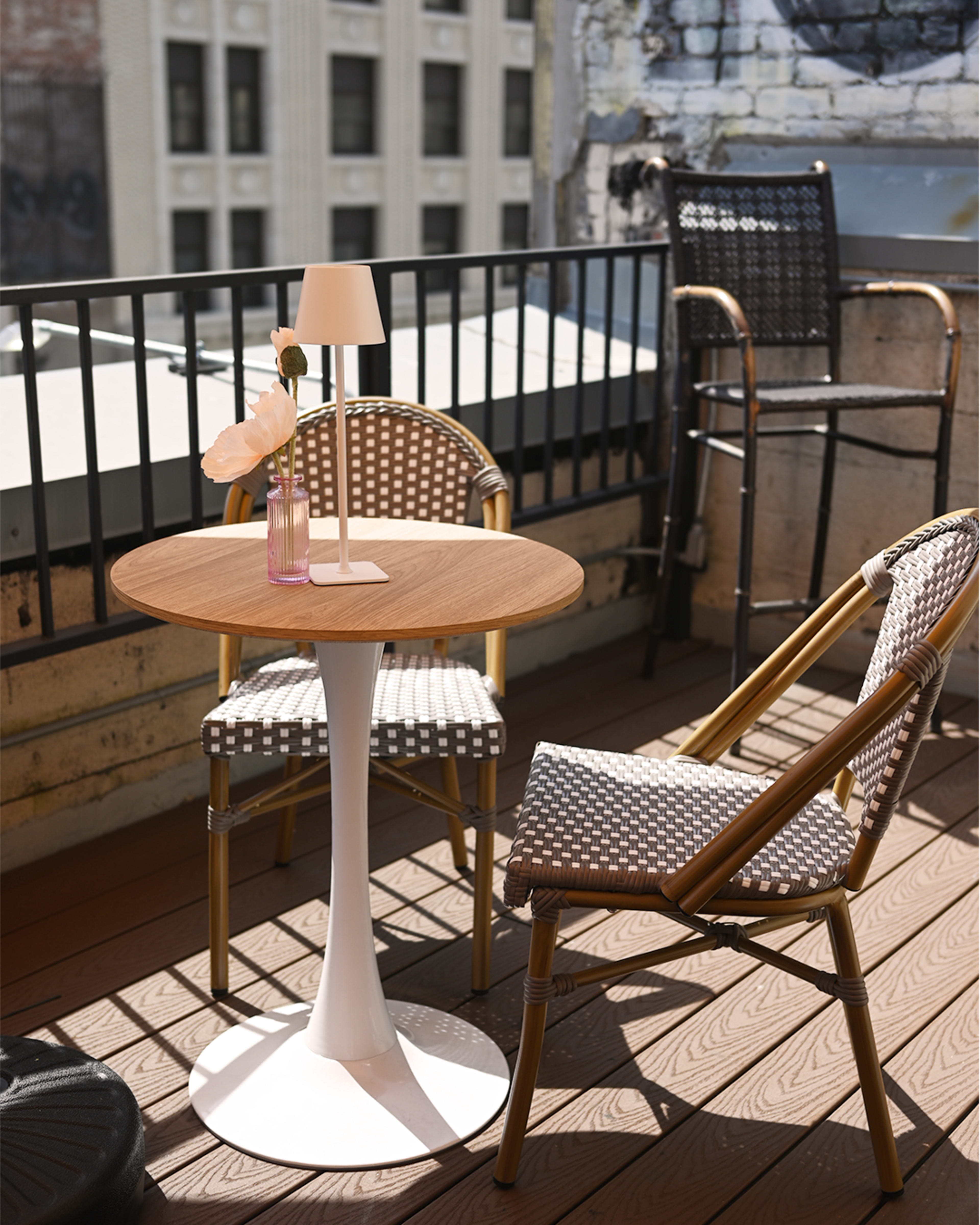 A small round table with a white base is set on a balcony, accompanied by two chairs and a lamp, overlooking a cityscape.