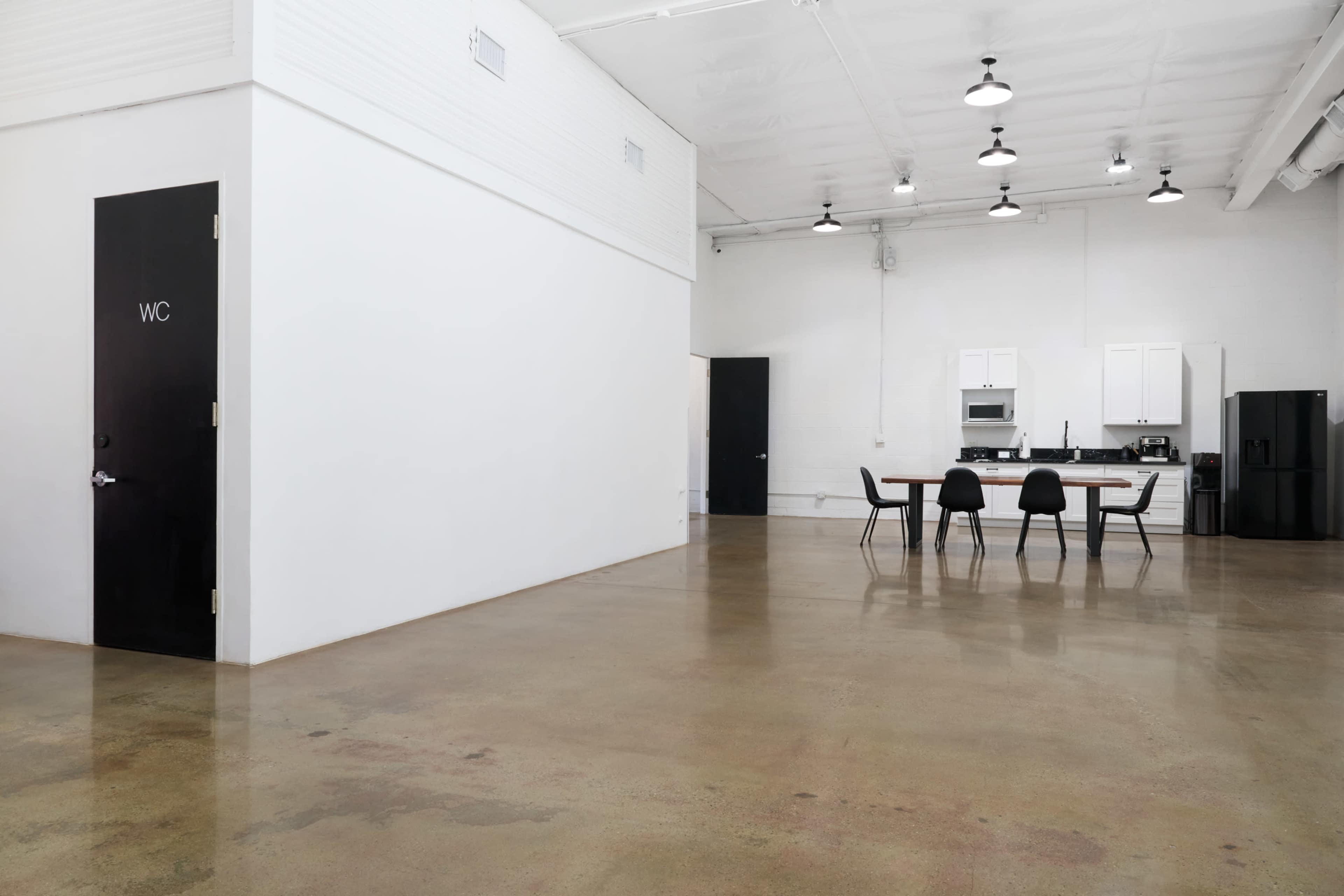 The image shows a spacious, modern industrial interior featuring a dining table with four chairs, a kitchen area, and a black door labeled "WC."