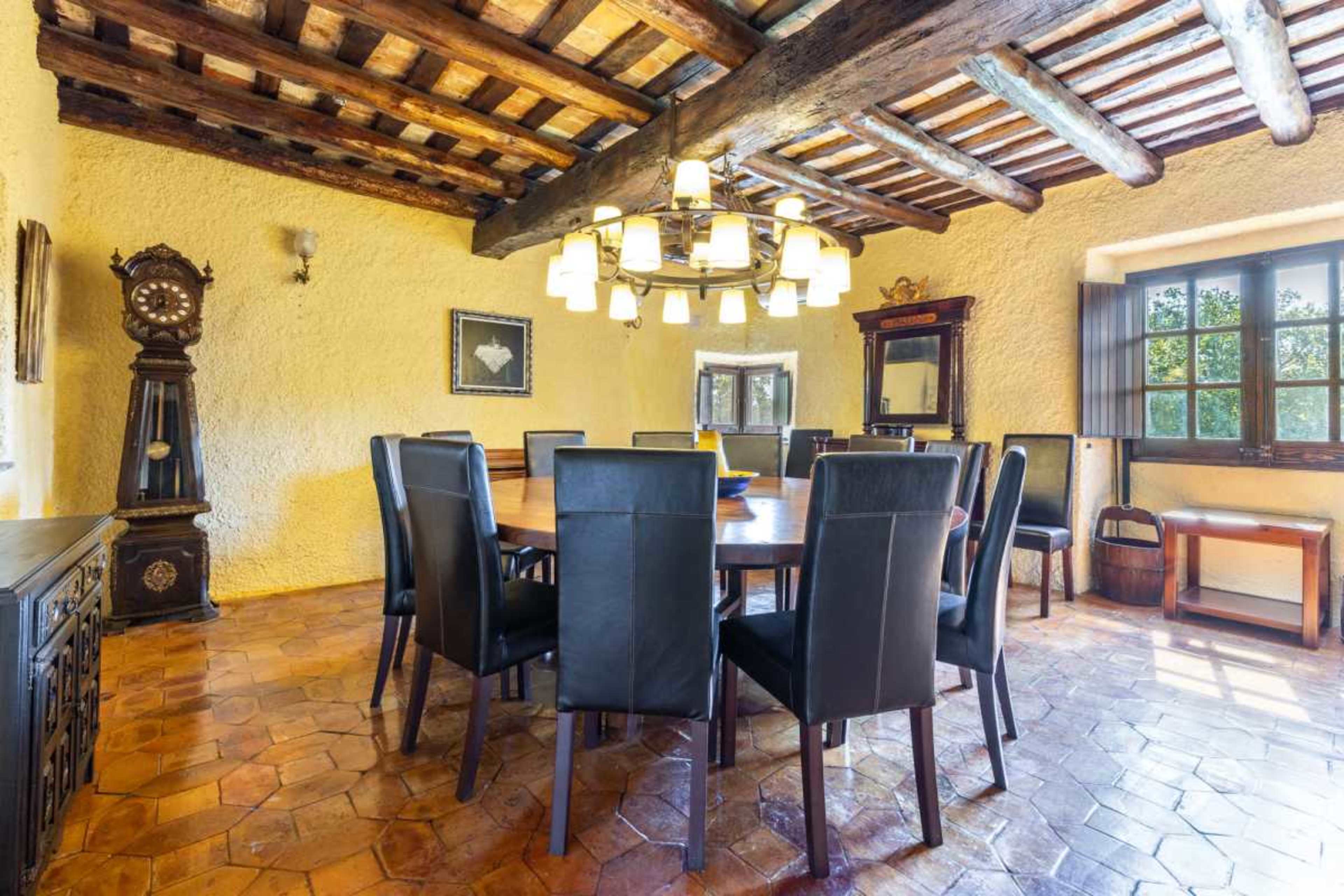 The image shows a rustic dining room featuring a large round wooden table surrounded by black chairs, with wooden beams on the ceiling and a grandfather clock in the corner.