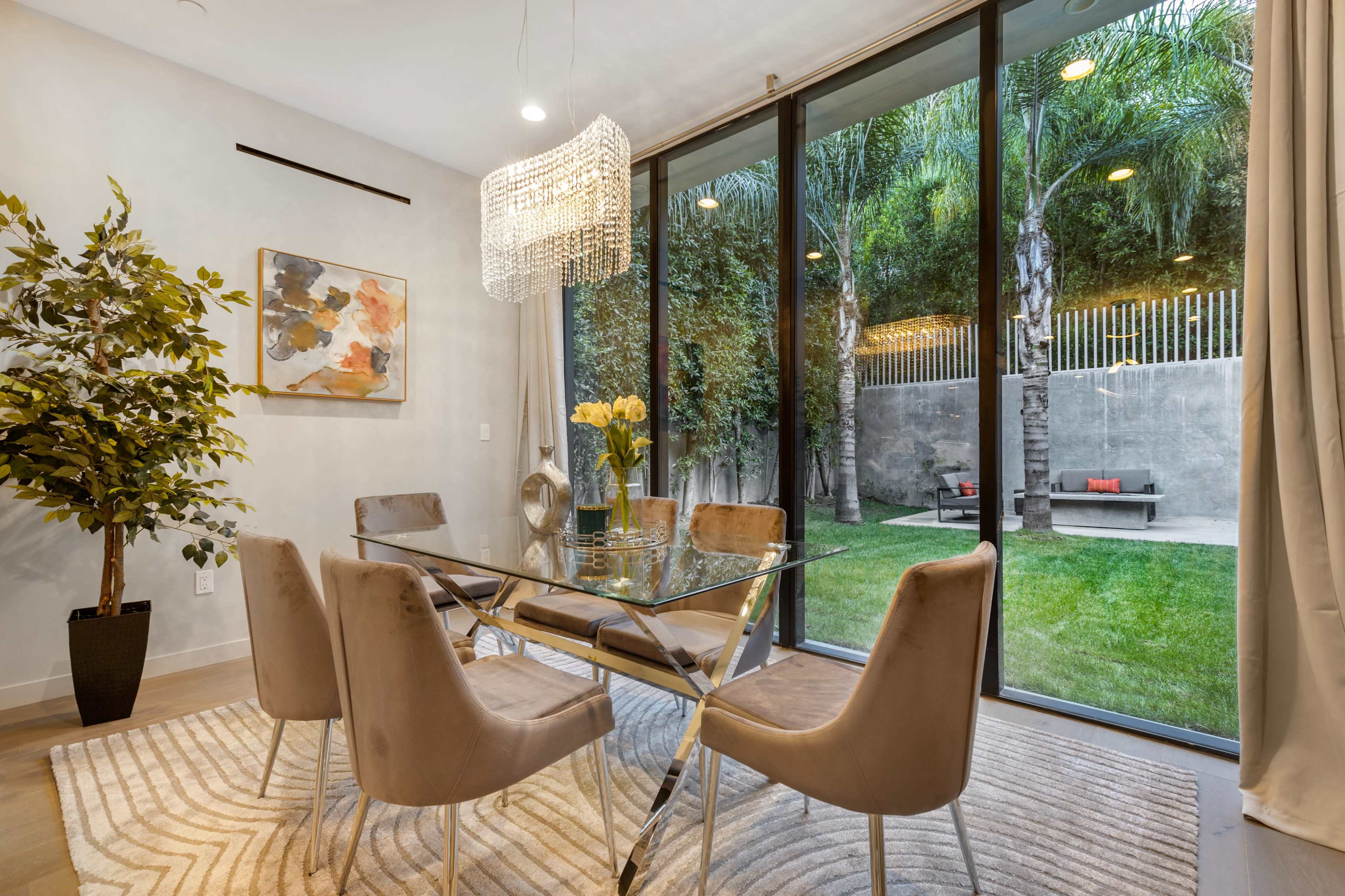 A dining area features a glass table surrounded by beige chairs, with a large window showcasing a green outdoor space.