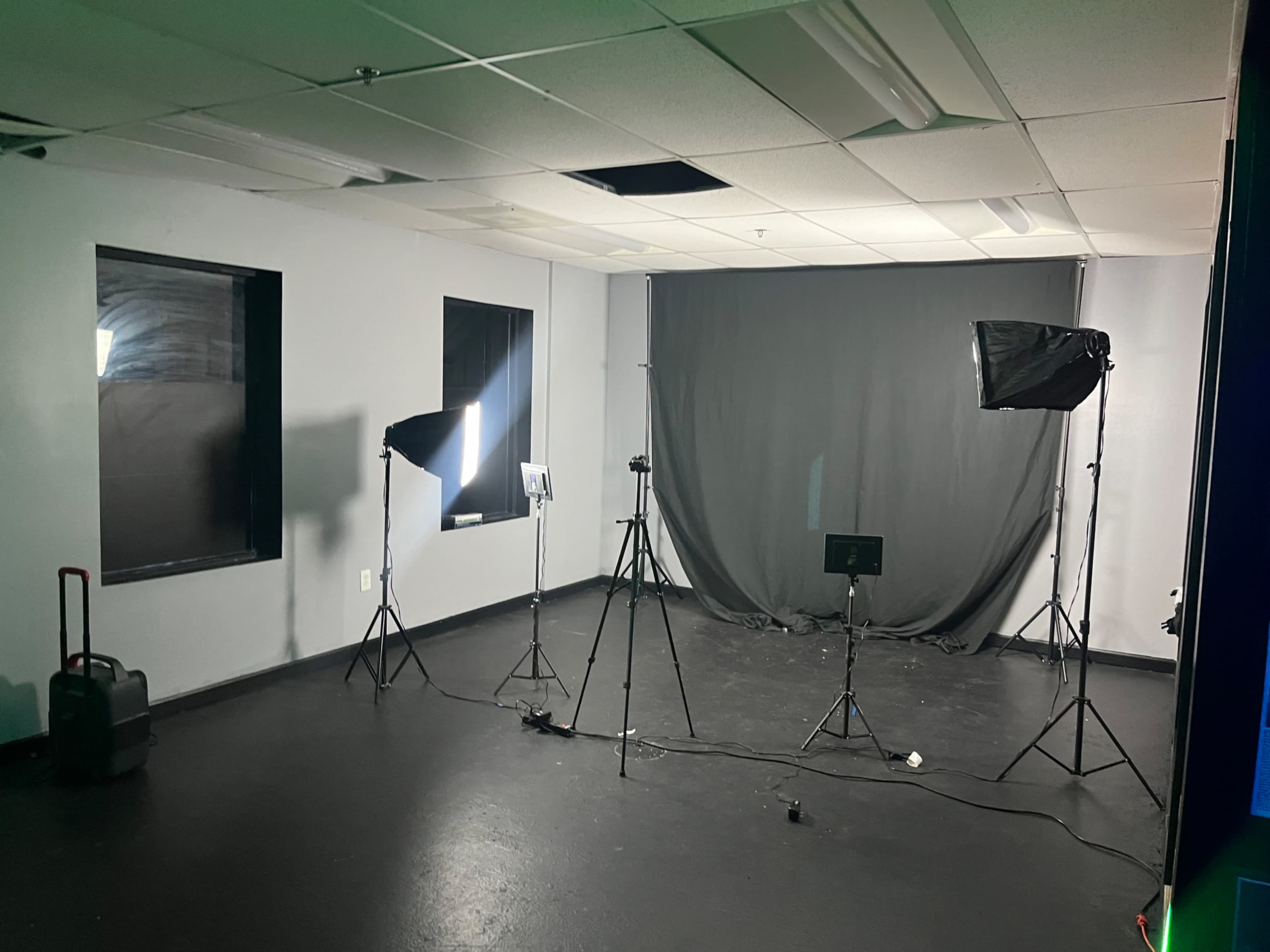 A room set up for video or photo shoots, featuring three tripods with lighting equipment, a gray backdrop, and a few pieces of portable gear.