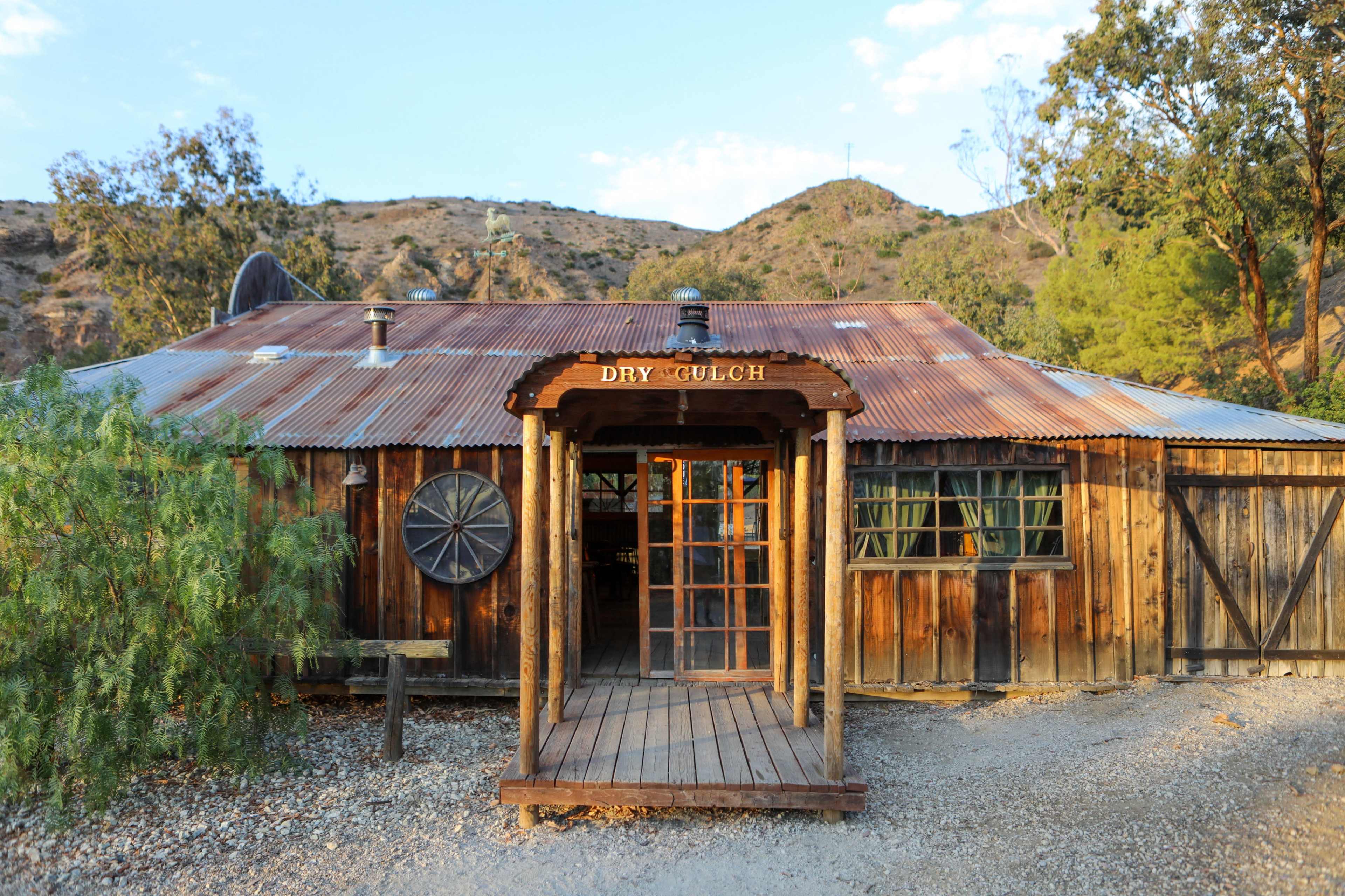 A rustic wooden building with a corrugated metal roof is situated on a gravel path, surrounded by trees and hills.
