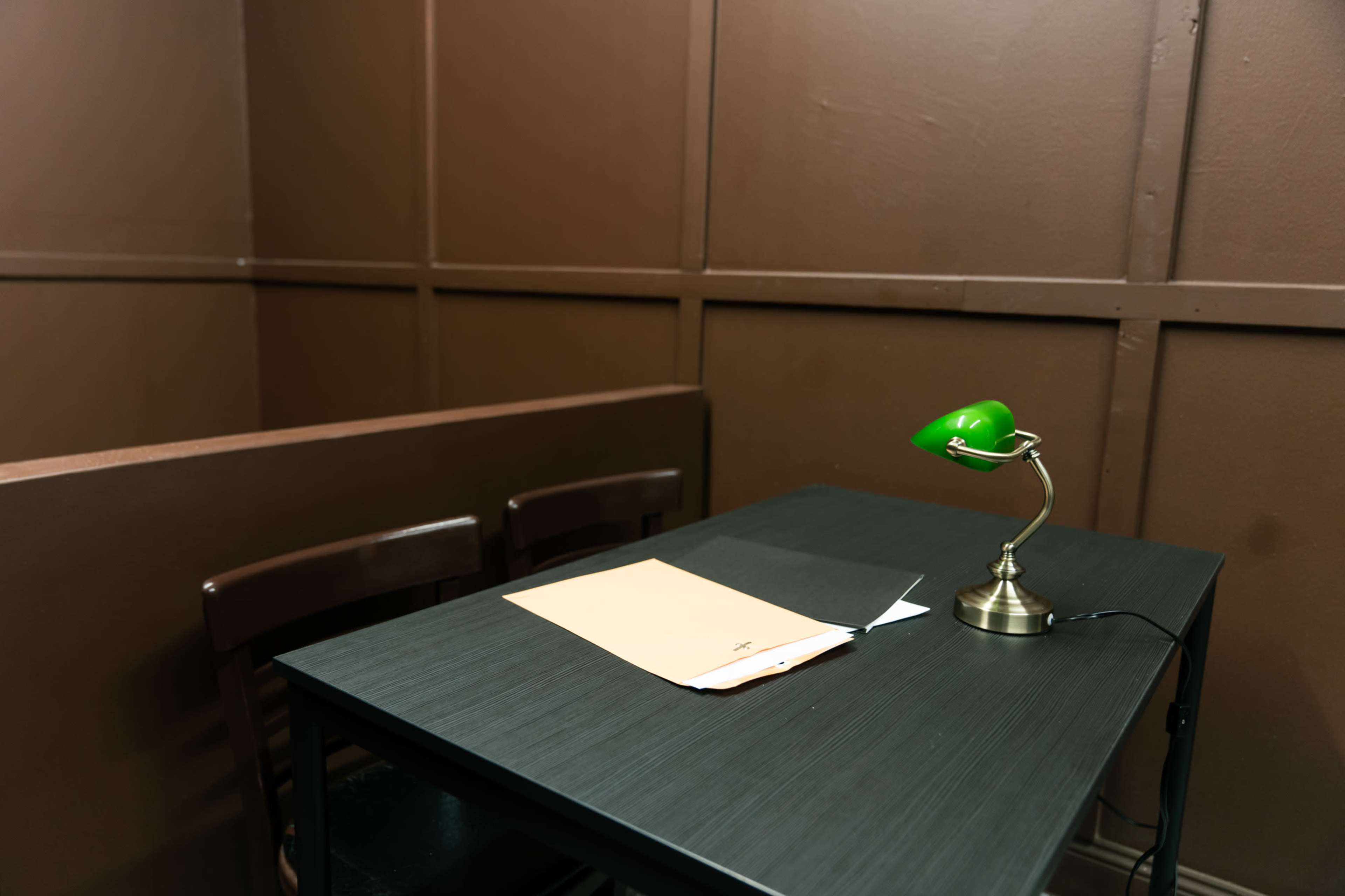 A small, dimly lit room features a black table with a green desk lamp and a folder on top, surrounded by brown wooden walls and chairs.