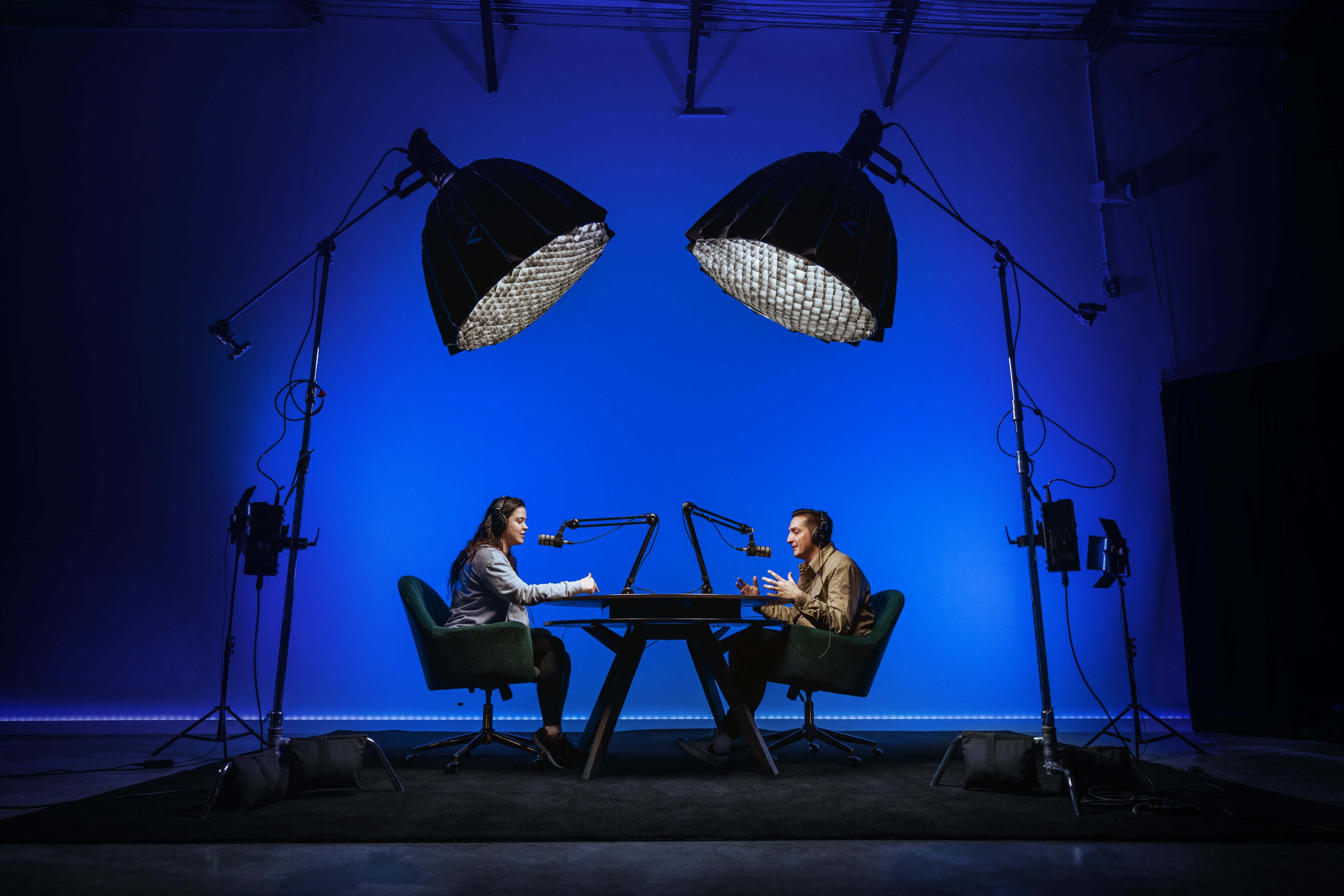Two people sit at a table in a studio setting, facing each other under large lights, with microphones positioned for a conversation against a blue backdrop.