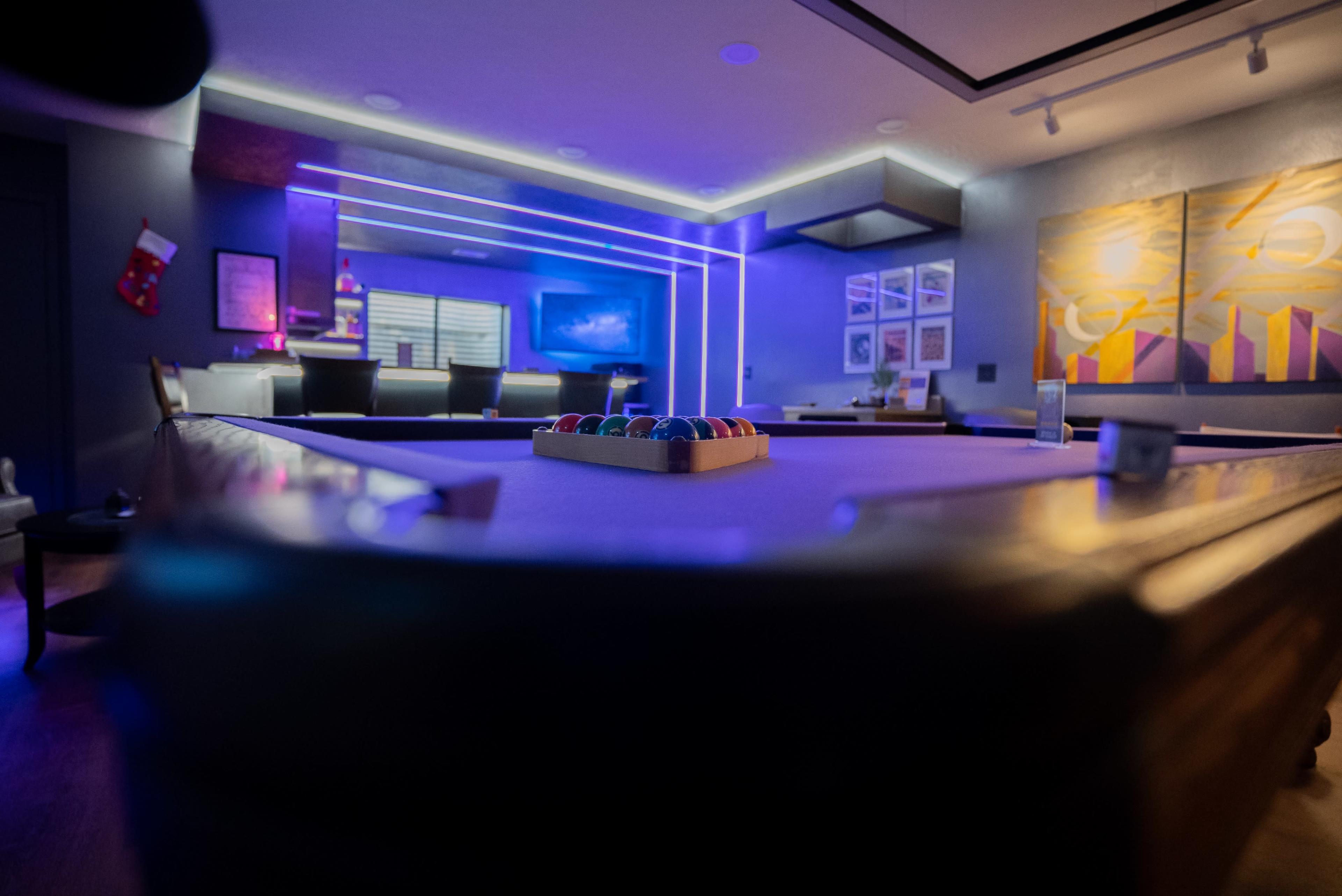 The image shows a modern game room with a pool table in the foreground, colorful lighting, and artwork on the walls.