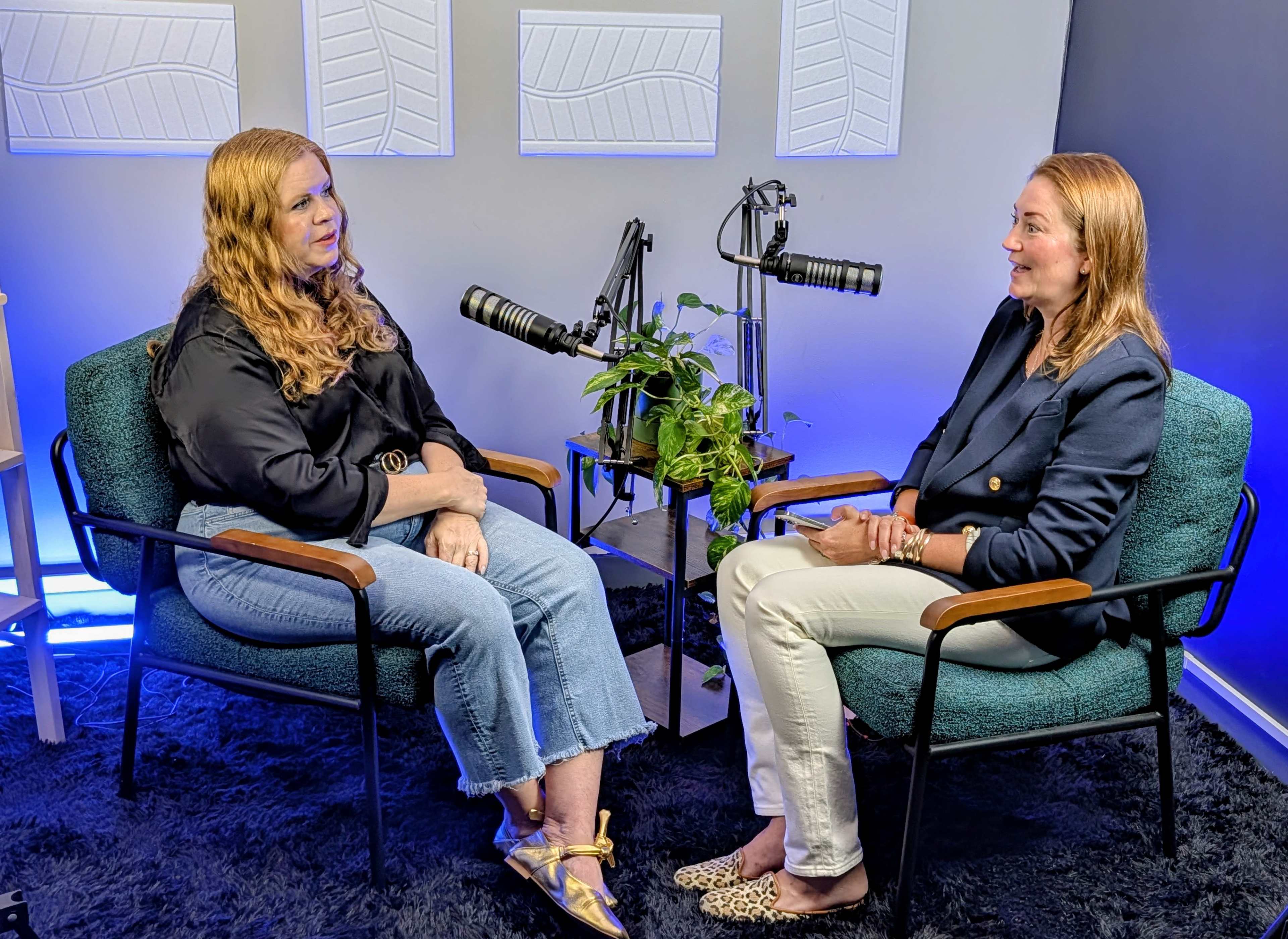 Two women are seated across from each other in a studio setting, with microphones positioned in front of them and a small plant on a table between them.