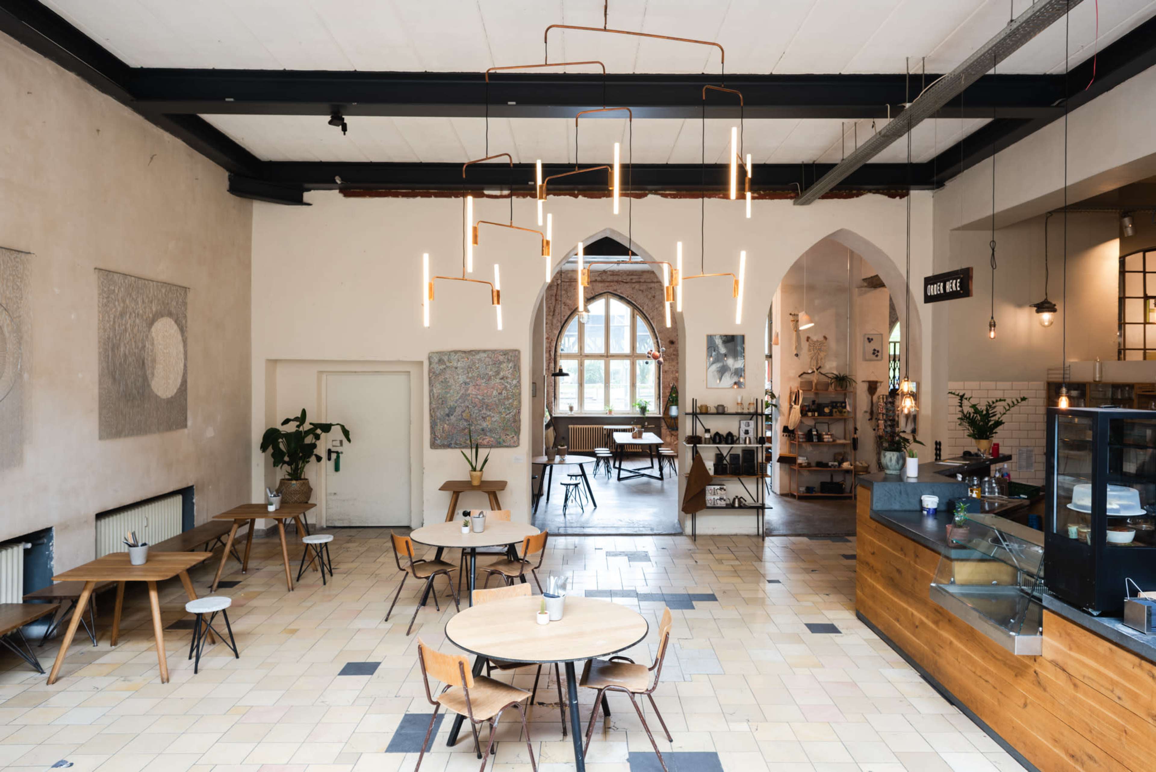 A spacious cafe with wooden tables and chairs, large windows, and modern light fixtures hanging from the ceiling.