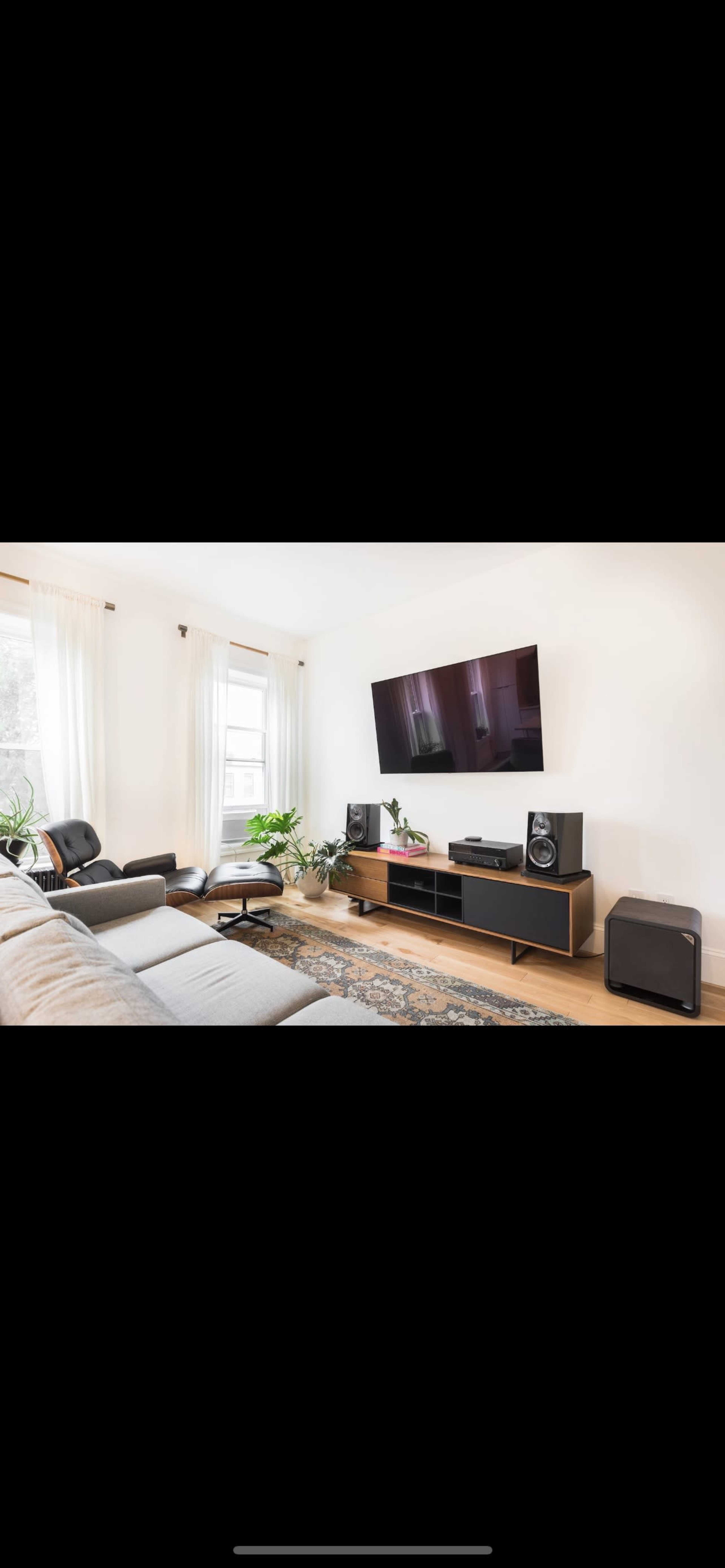 A modern living room features a gray sectional sofa, wooden media console with speakers, a large TV mounted on the wall, and several indoor plants.