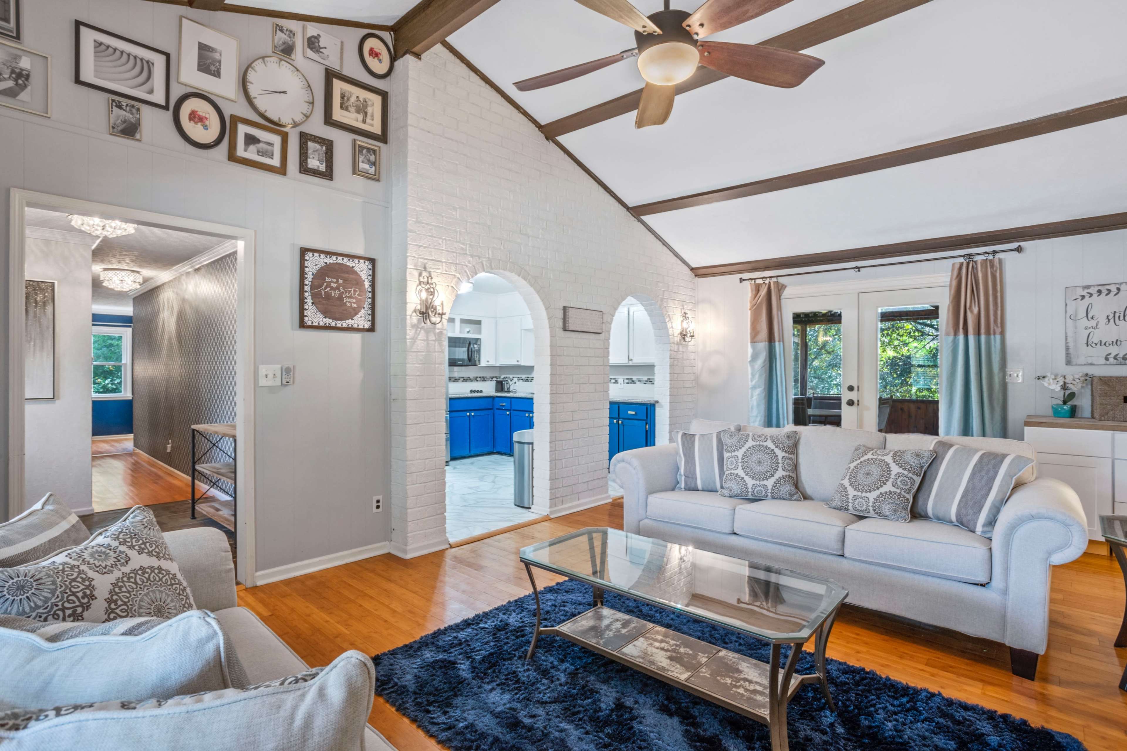 A living room features a light-colored sofa set, a glass coffee table on a dark rug, and a vaulted ceiling with exposed beams, leading to an adjoining hallway and kitchen area.