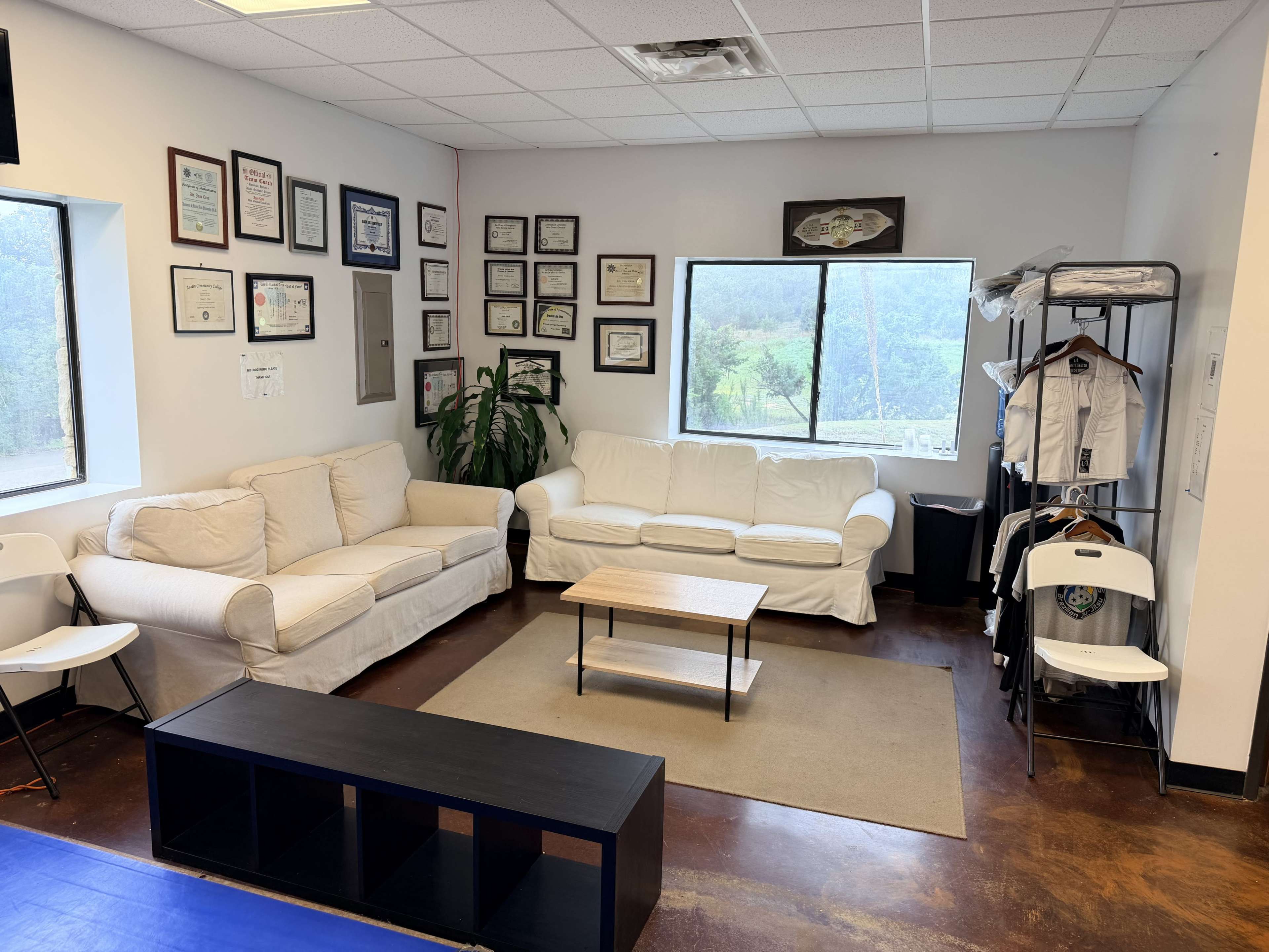 The image shows a waiting area with two white sofas, a small coffee table, a large window providing a view of greenery, and framed certificates on the wall.