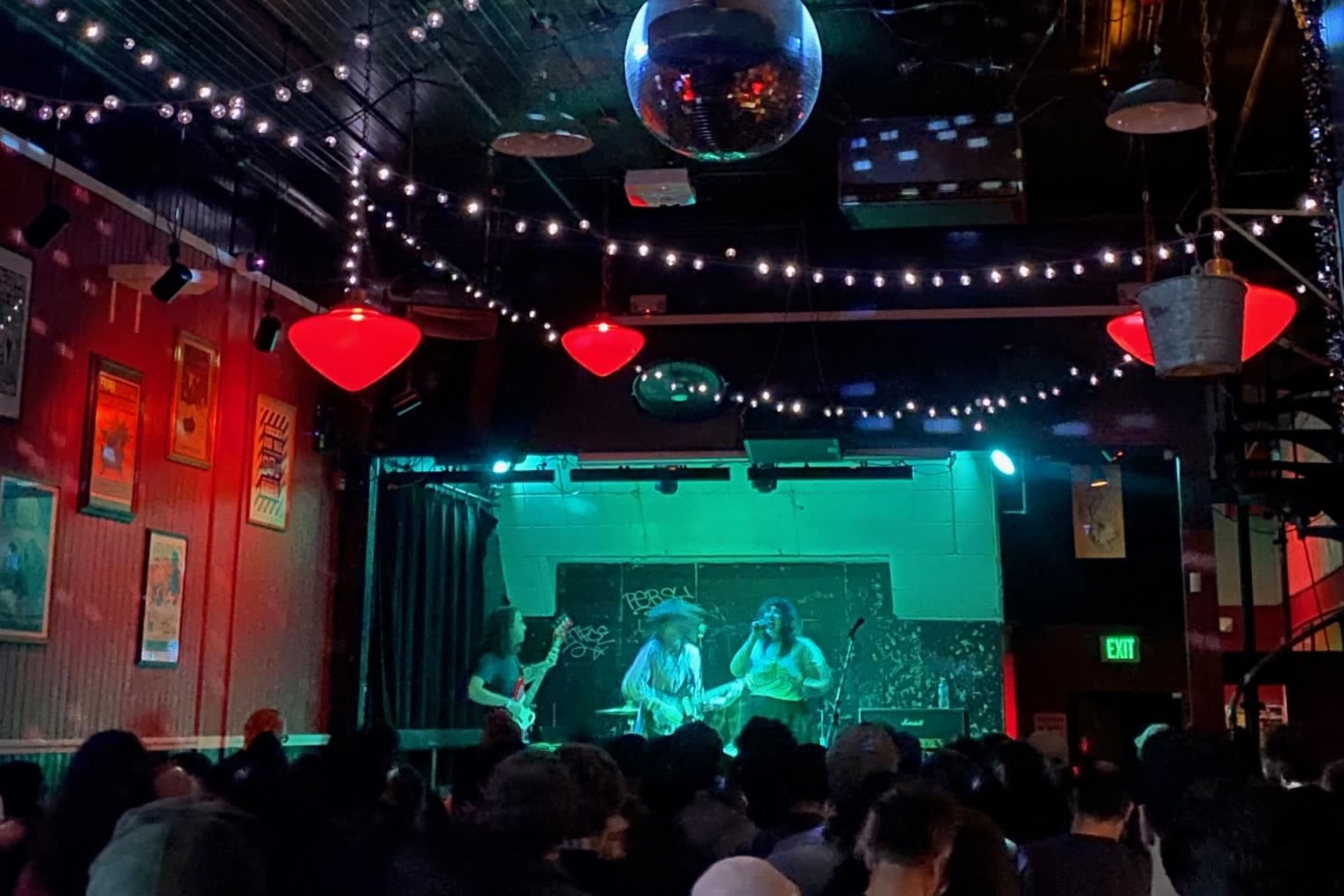 A band performs on stage in a dimly lit venue filled with a crowd, under glowing lights and a disco ball.