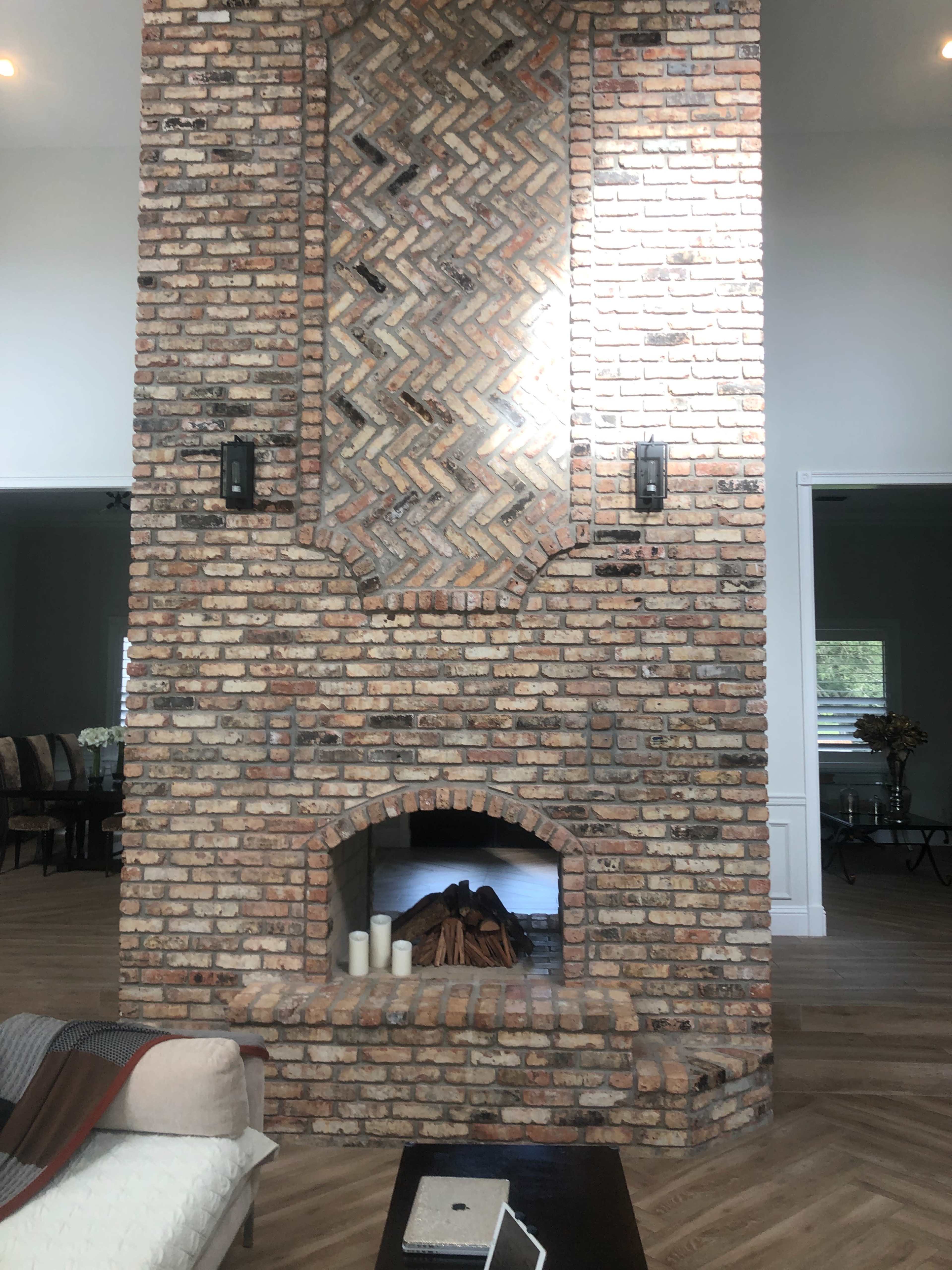 A tall, textured brick fireplace with an arched opening and two wall sconces, set against a light-filled room with wood flooring.